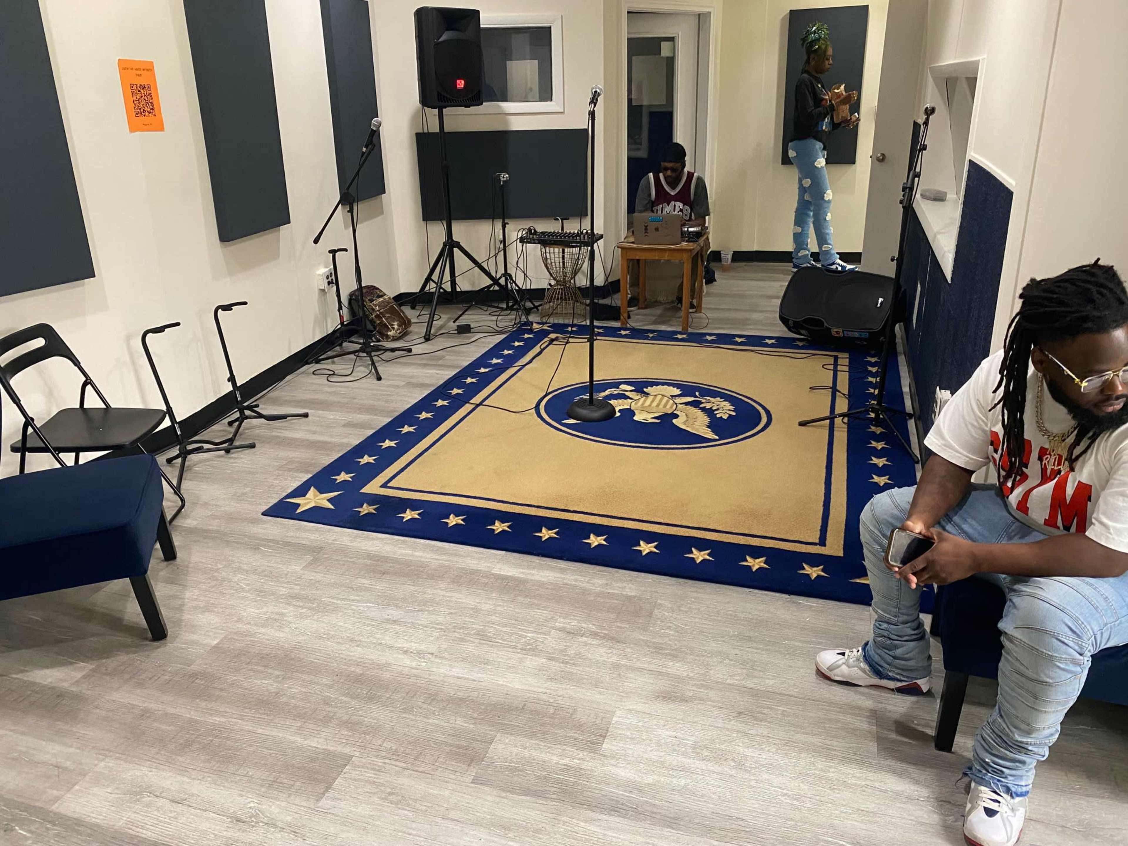 A rehearsal room with a microphone setup, musical equipment, and two people present, one seated and another working at a table.