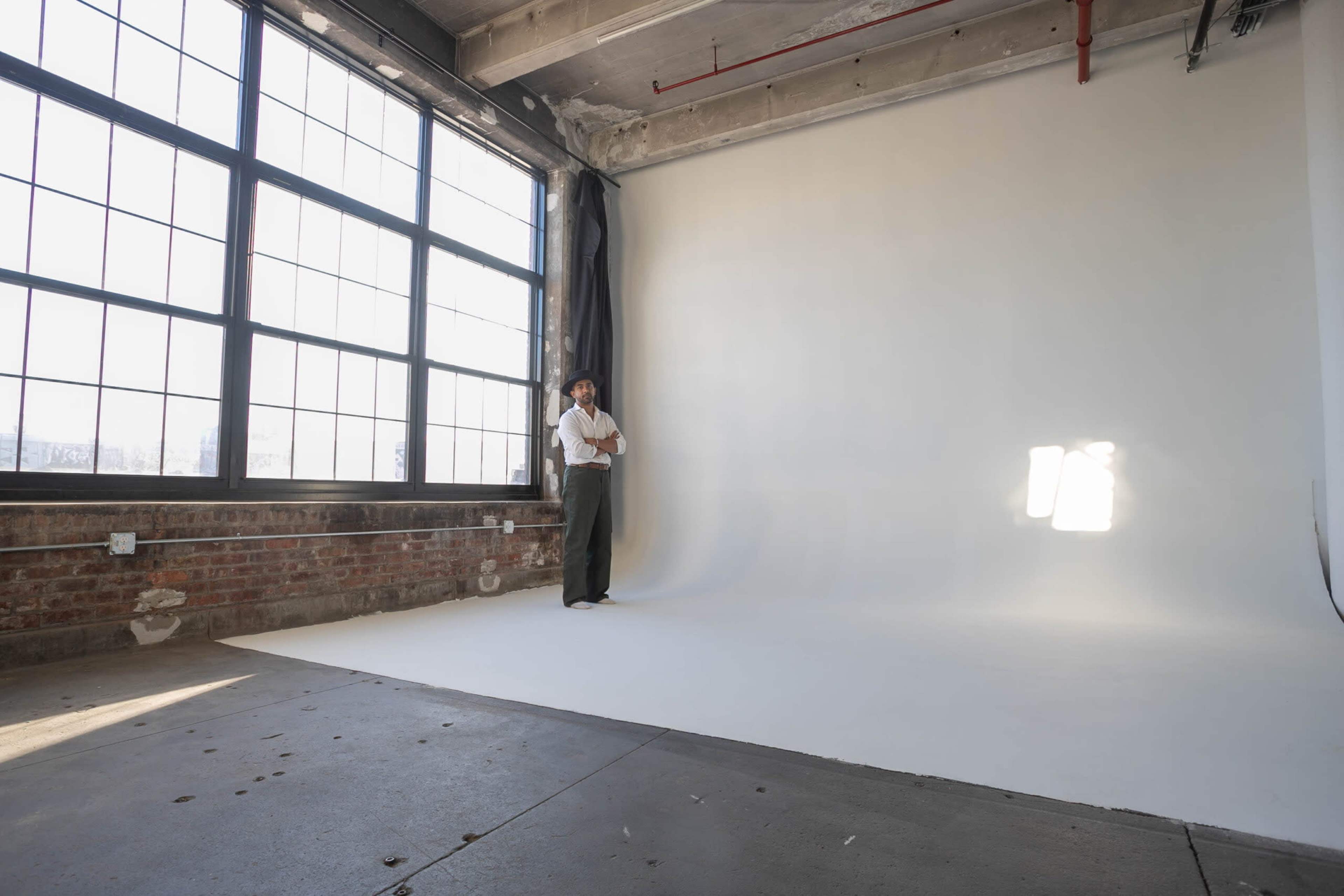 A person stands in a large, empty studio featuring a white backdrop and large windows allowing natural light to stream in.
