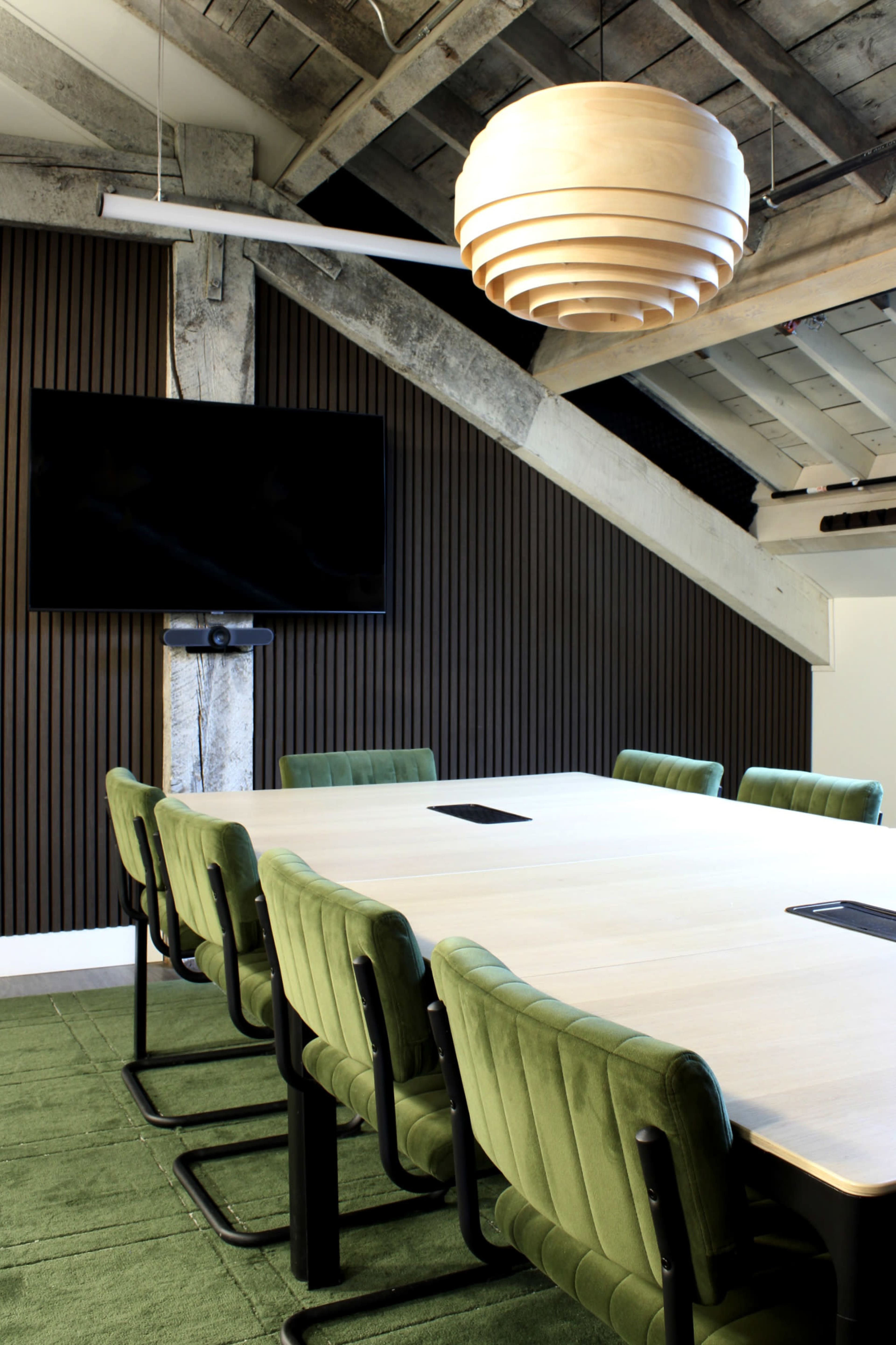 A conference room features a large table surrounded by green chairs, with a wooden pendant light hanging above and a television mounted on the wall.