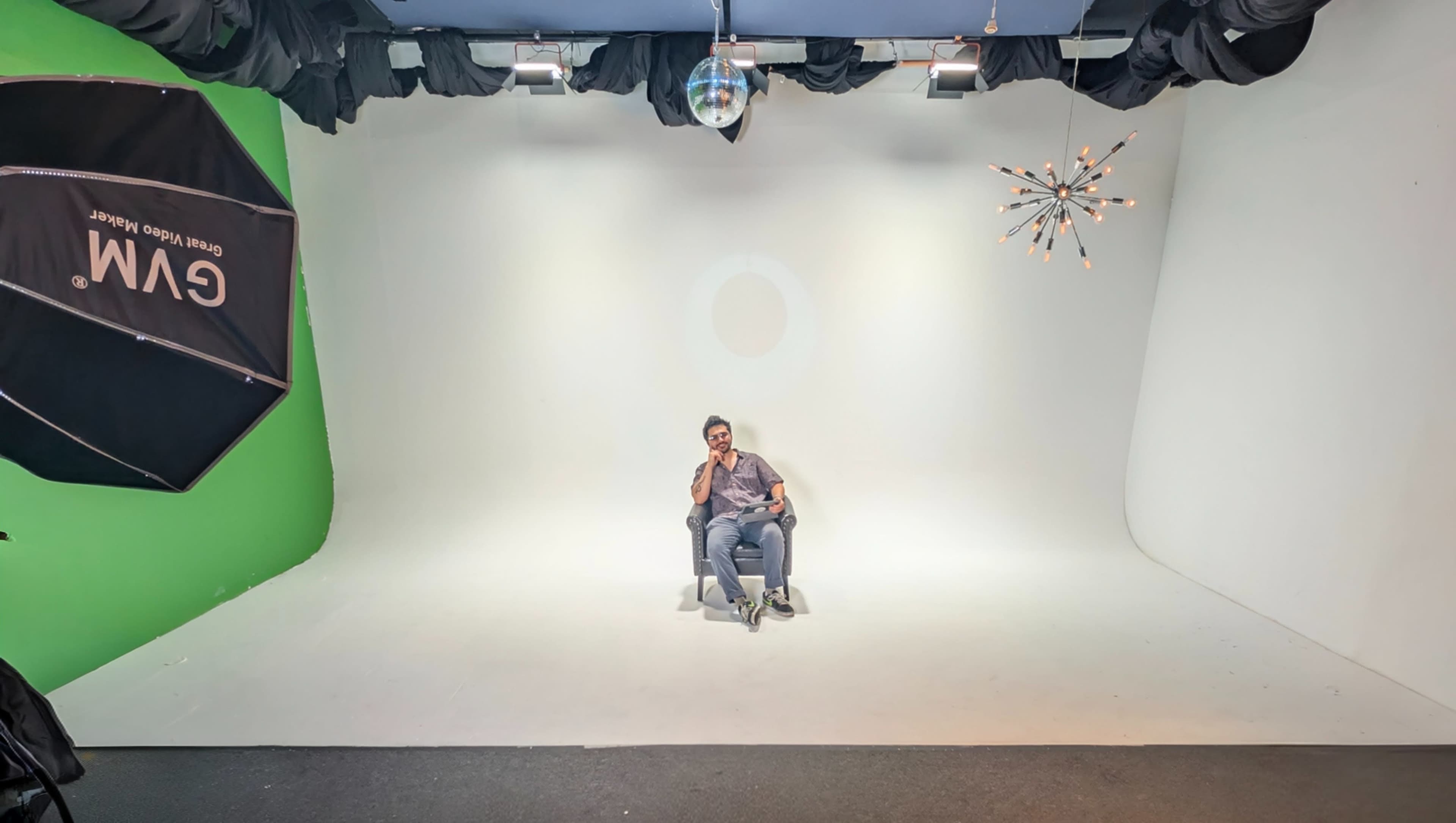 A person is seated on a chair in a studio with a white backdrop, a green screen, and lighting equipment.