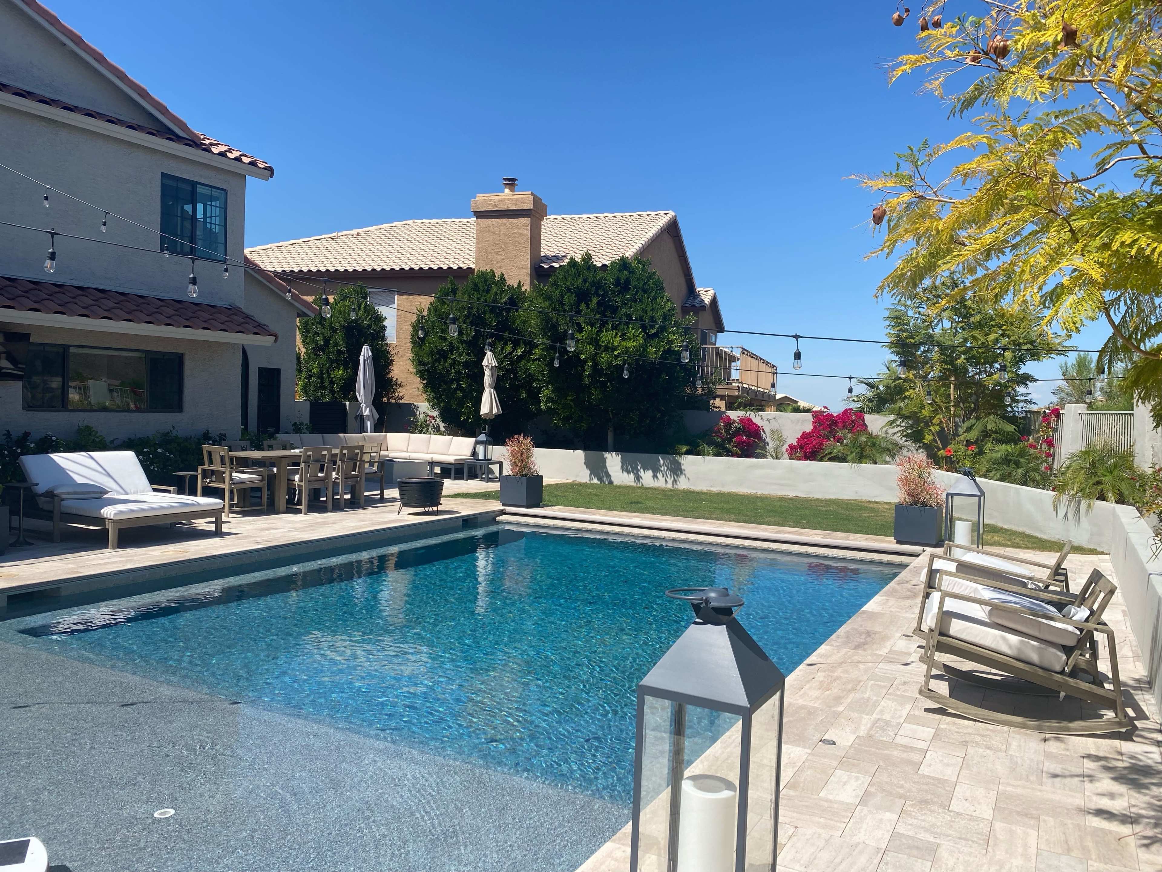 A residential backyard featuring a swimming pool, lounge chairs, a dining area, and landscaped greenery against a clear blue sky.