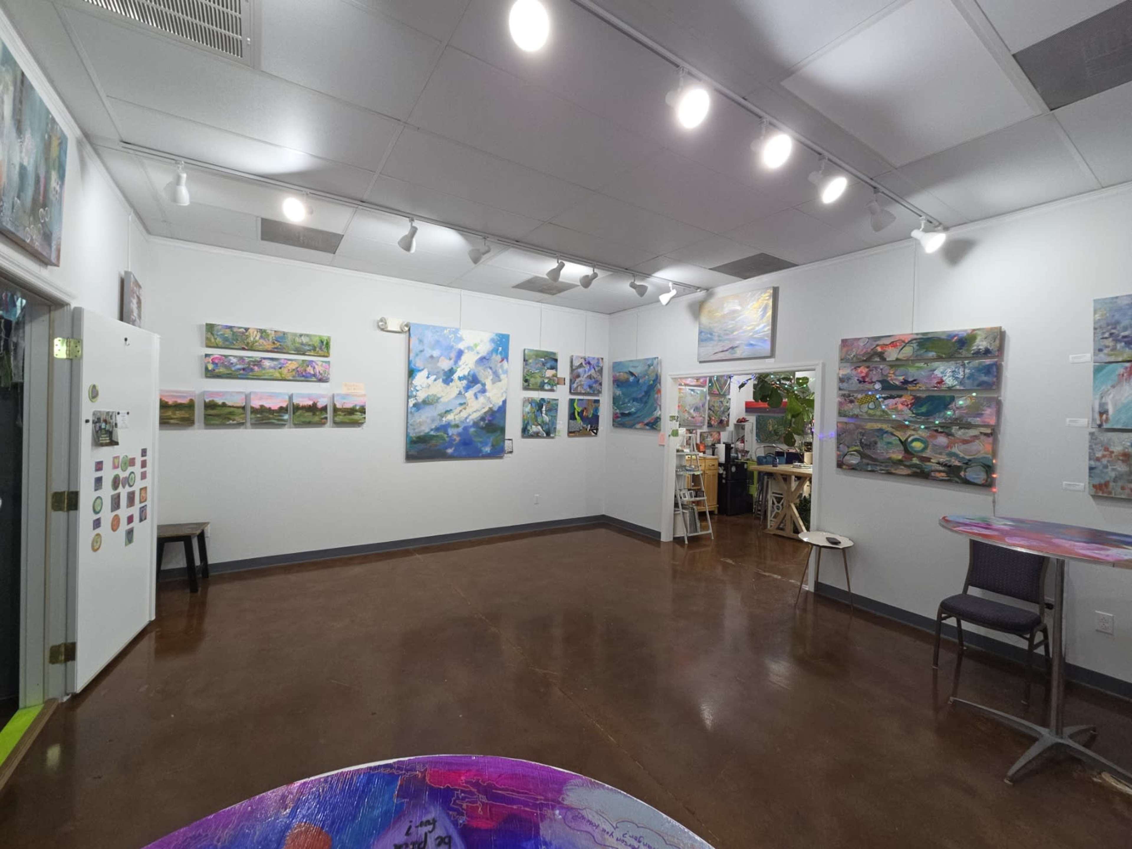 The image shows an art studio with colorful abstract paintings displayed on white walls and two tables in the foreground.