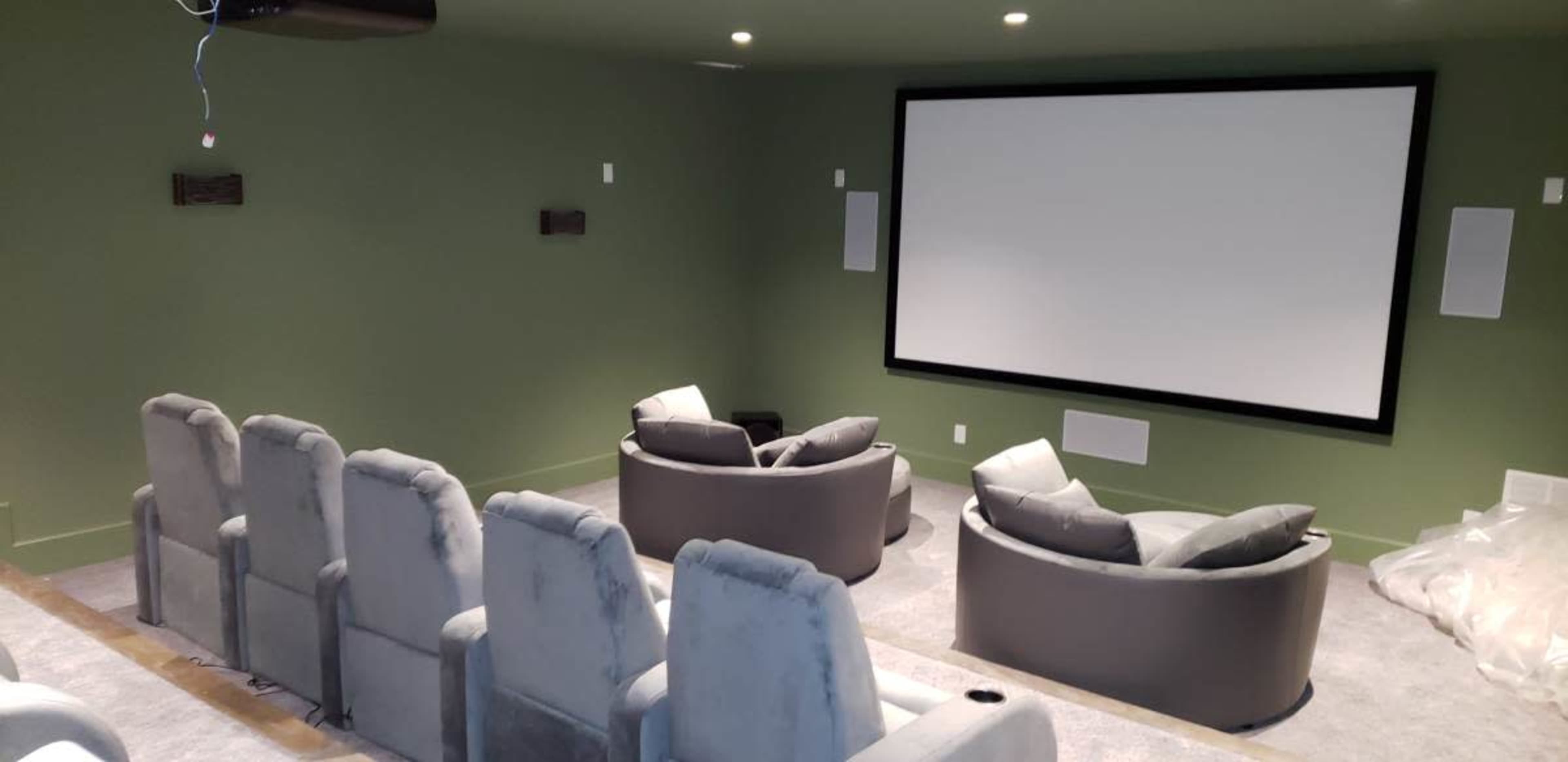 A home theater room features gray upholstered seating arranged in front of a large blank screen on a green-painted wall.