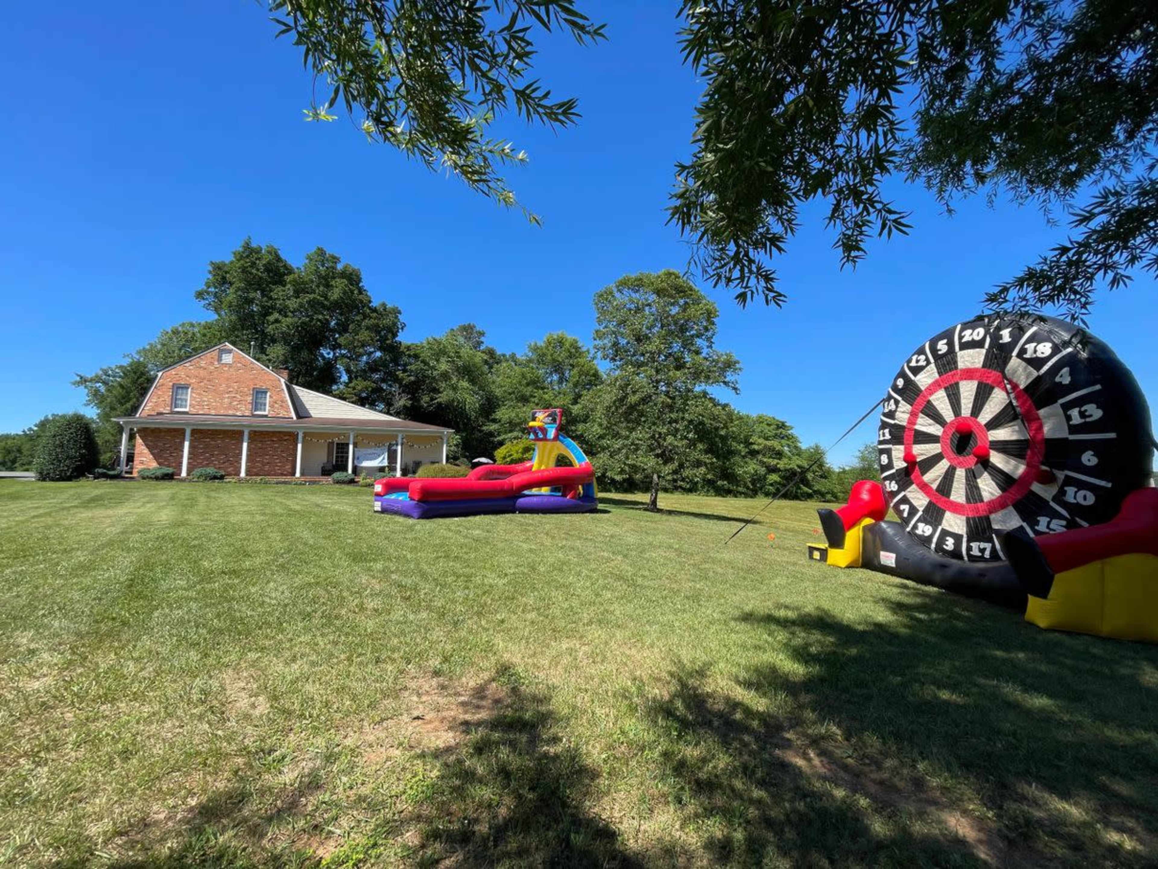 A large inflatable dartboard and a colorful bounce house are set up on the lawn of a brick house on a clear day.