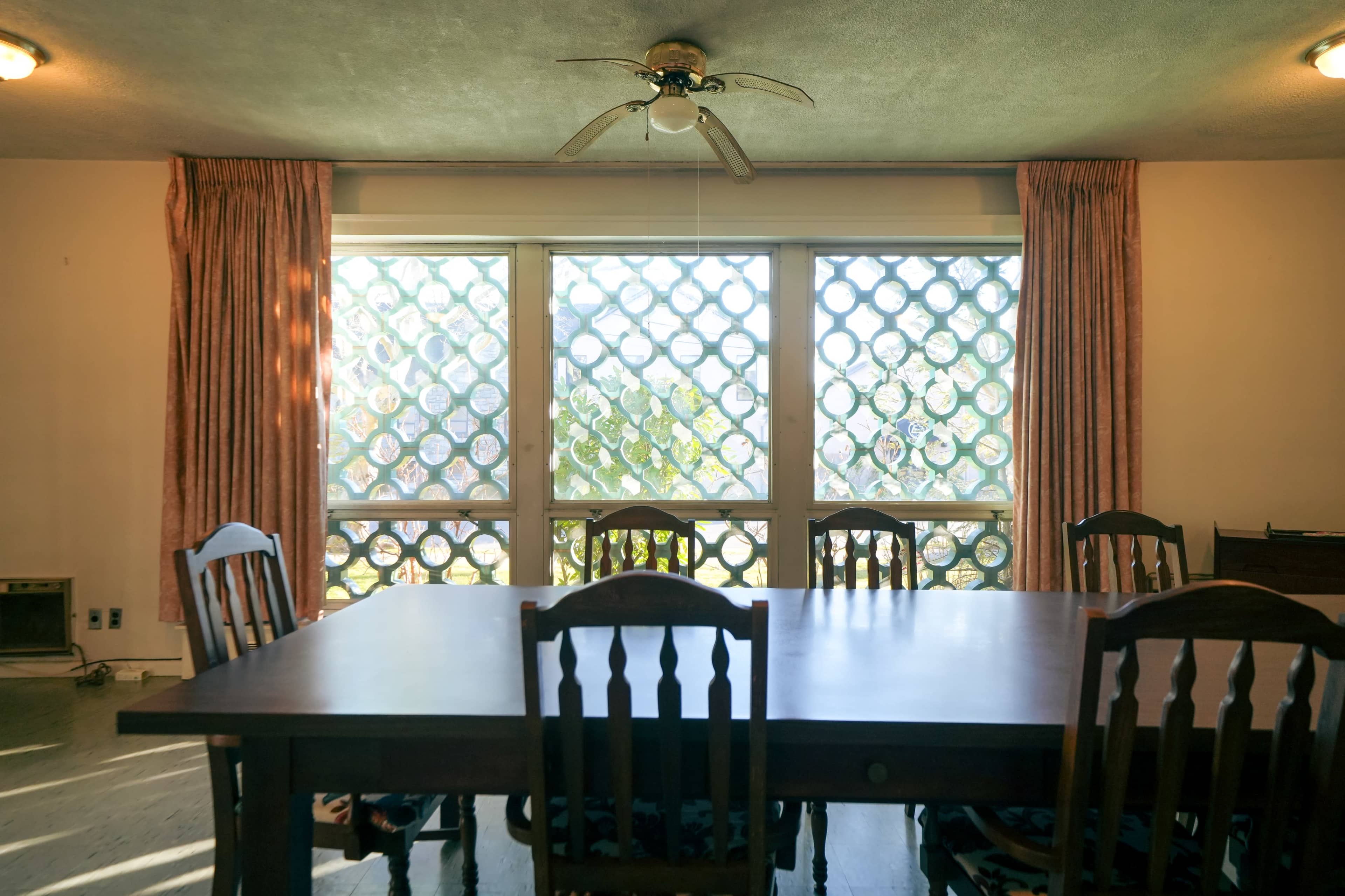 A wooden dining table with several chairs is positioned in front of large windows adorned with decorative lattice panels.