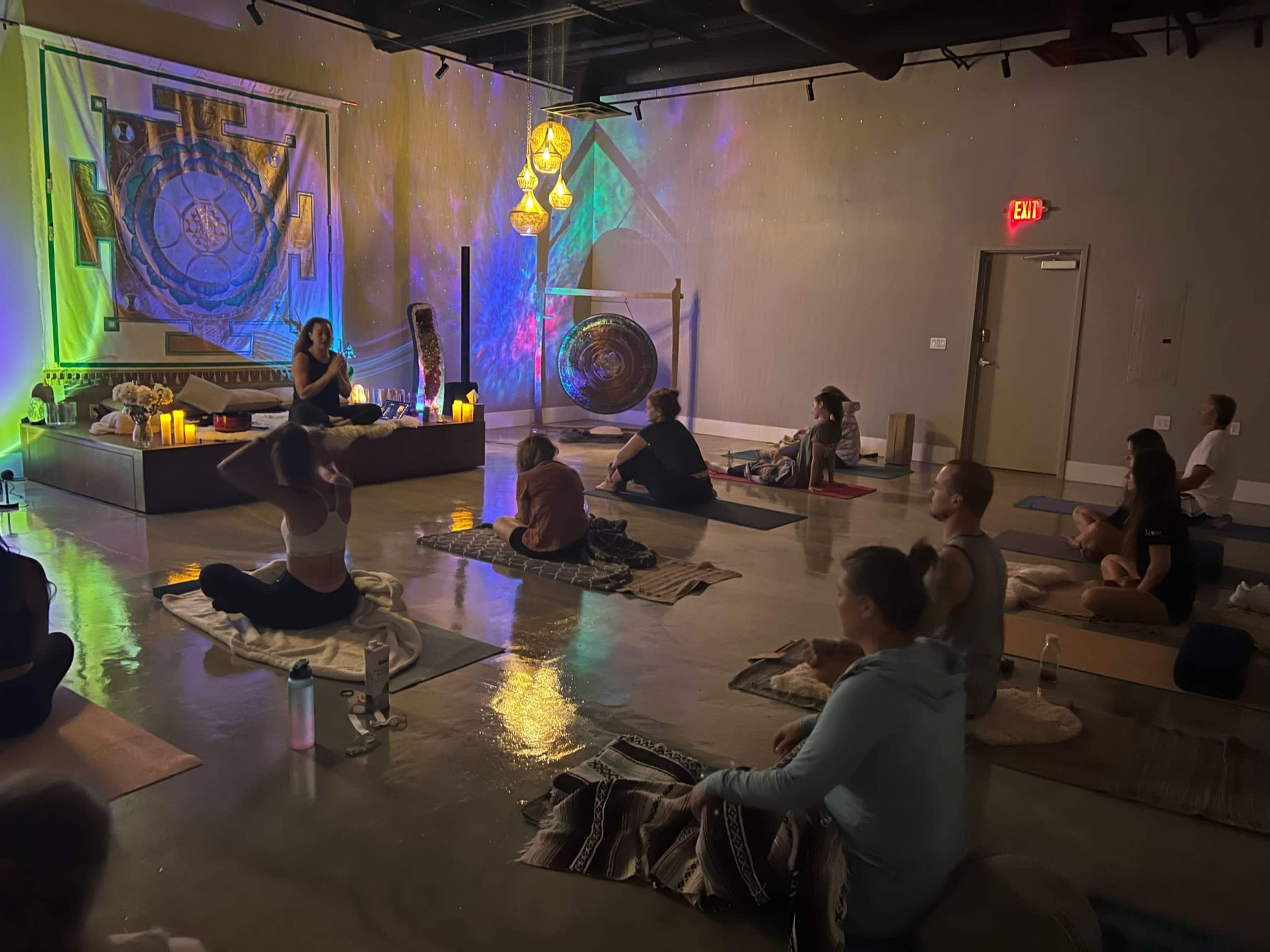 A group of individuals sits on yoga mats in a dimly lit studio, while a person leading the session is positioned at the front near candles and a large gong.