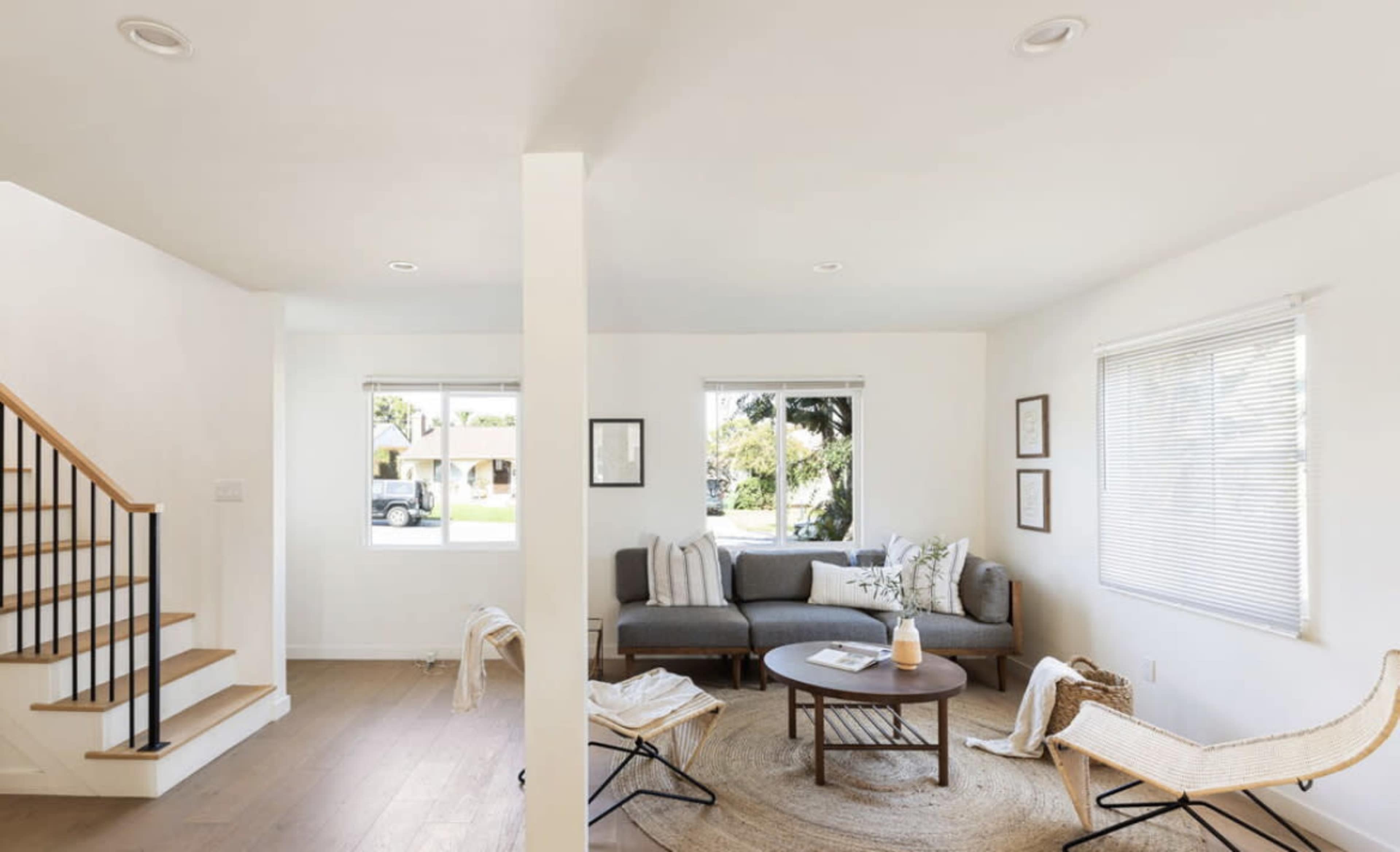 The image shows a bright, modern living room with a gray sofa, a round coffee table, and large windows allowing natural light to enter.