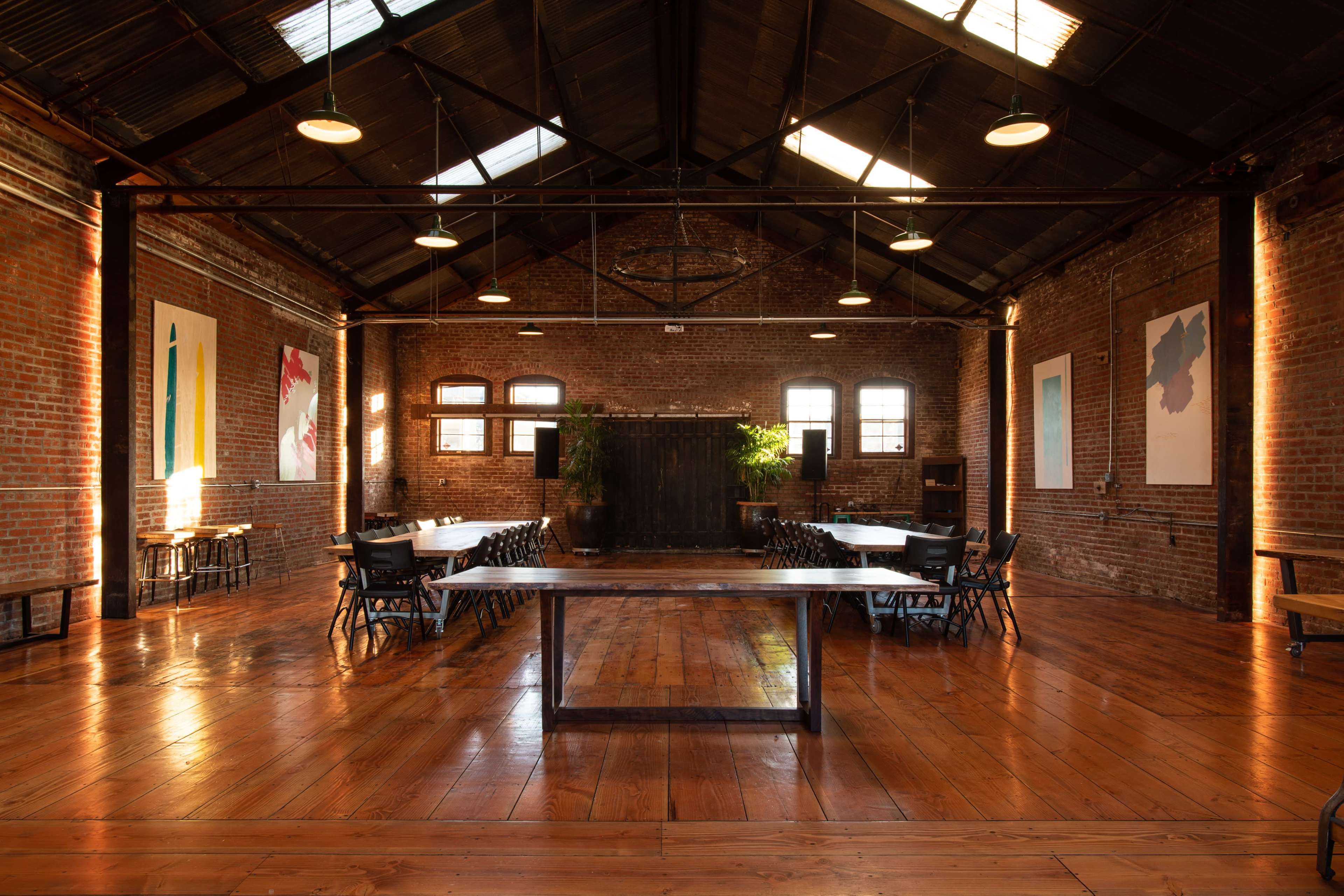 A spacious industrial-style room with brick walls, large windows, and a table set up for a meeting or event.