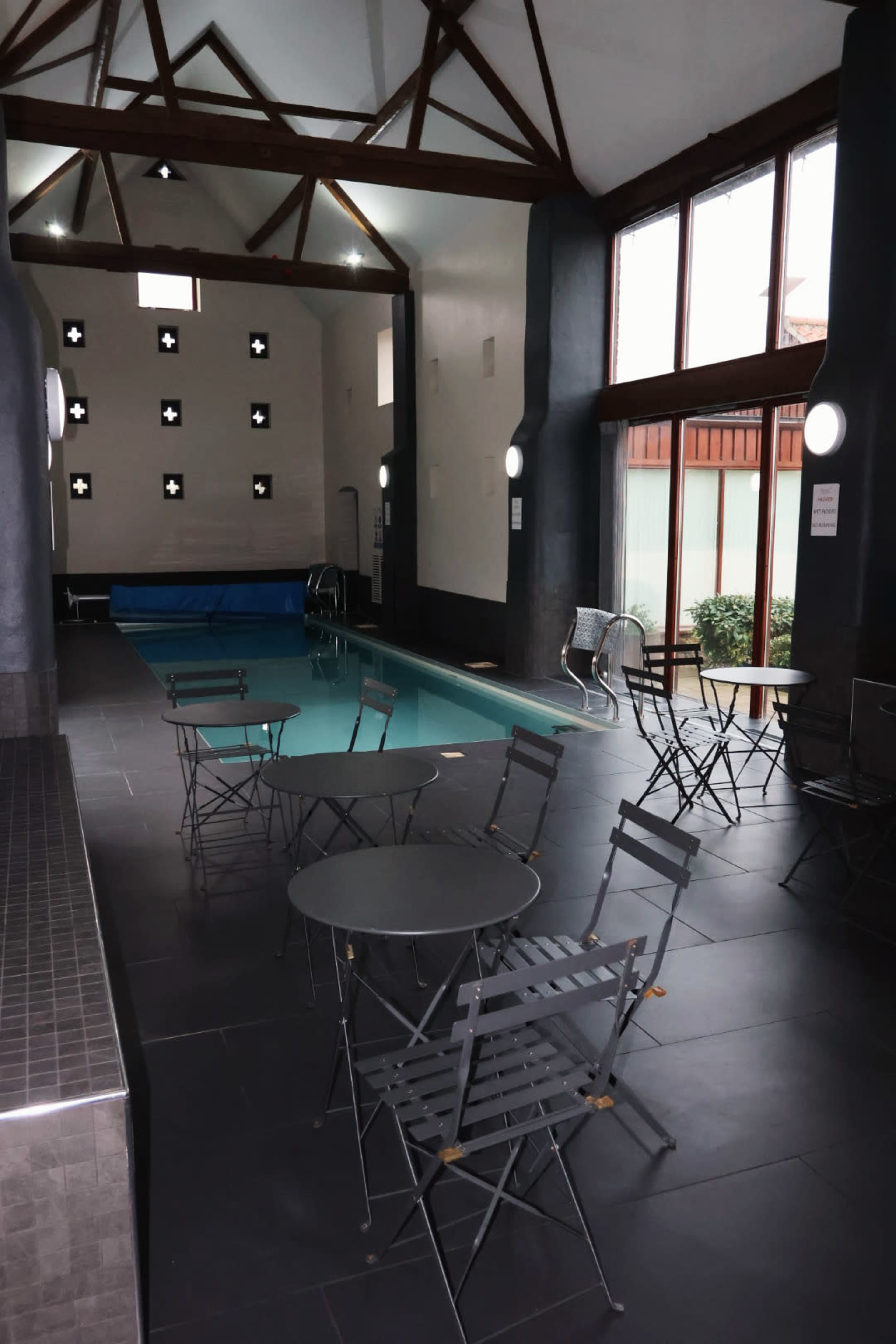 The image shows an indoor swimming pool area with a small pool, grey folding chairs, and large windows letting in natural light.