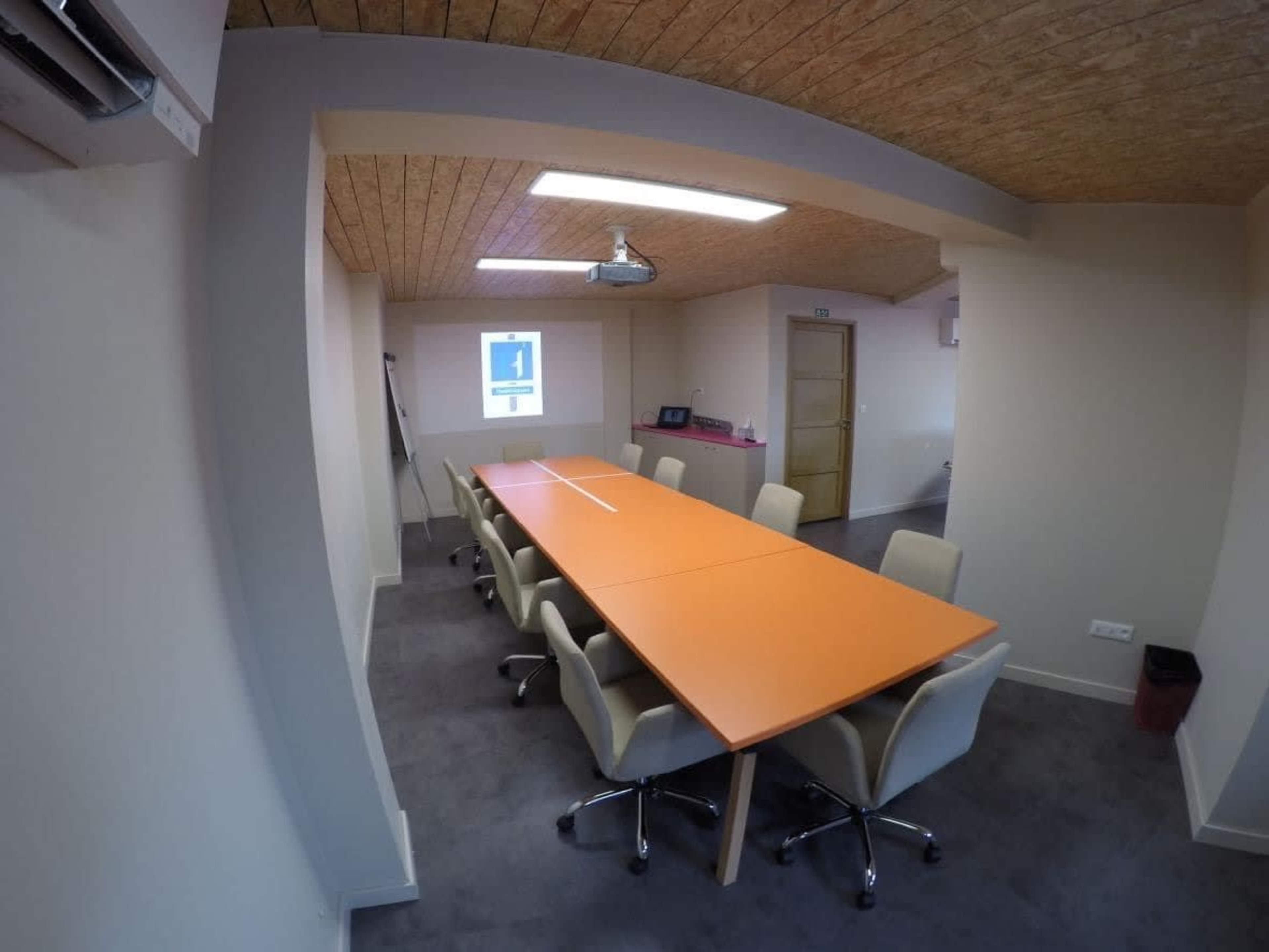The image shows a conference room with a long orange table surrounded by light-colored chairs and a projector mounted on the ceiling.