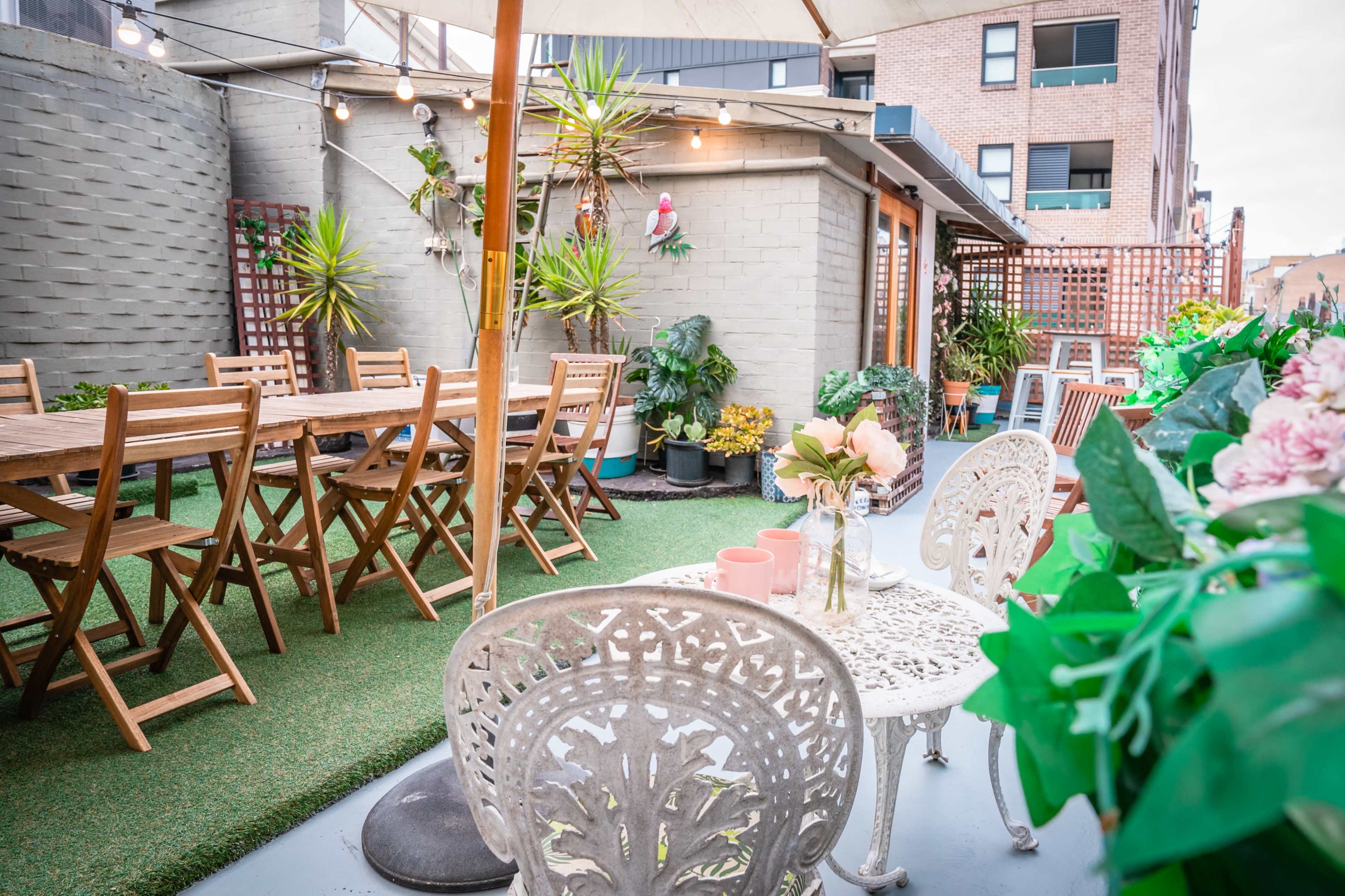 A rooftop garden with wooden dining tables, potted plants, and decorative chairs surrounded by a simple, modern building.
