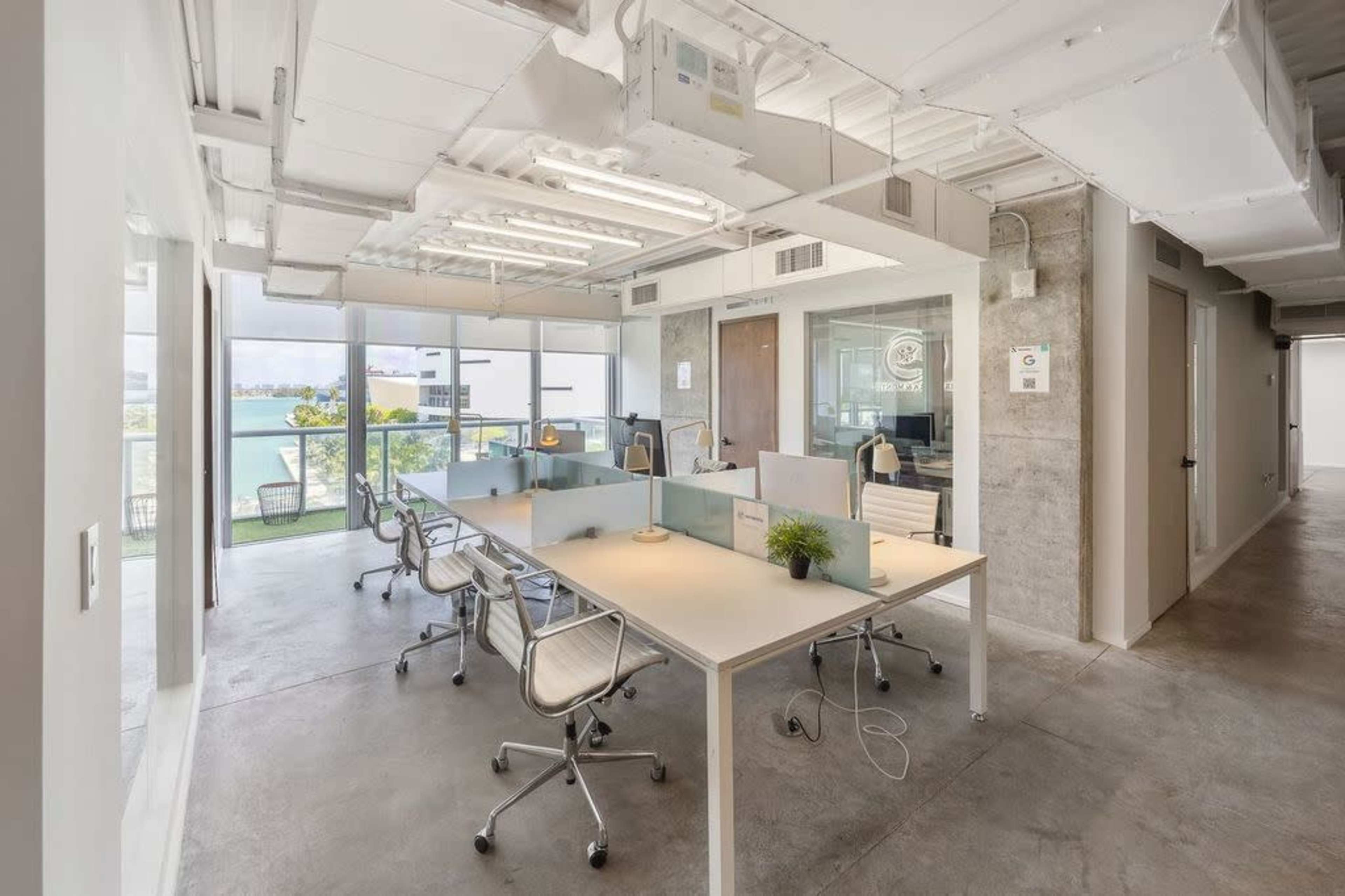 A modern office space with several workstations, large windows overlooking a waterfront, and minimalistic decor.