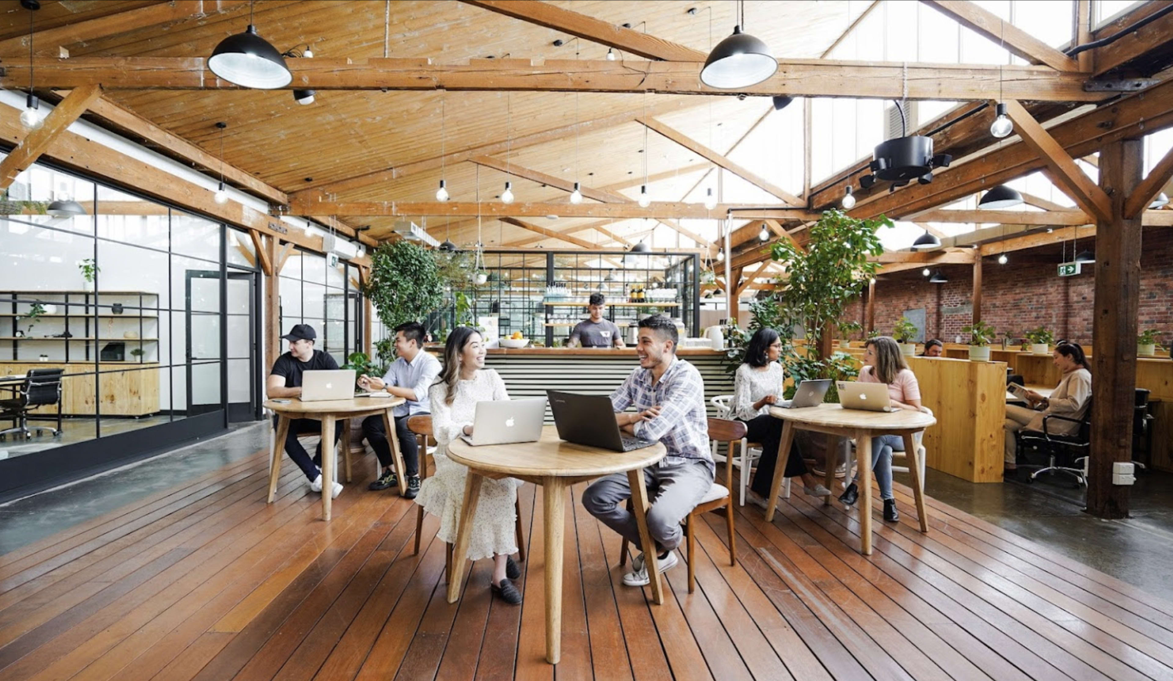A modern workspace features several individuals working on laptops at round wooden tables under a wooden beam ceiling with plants throughout.