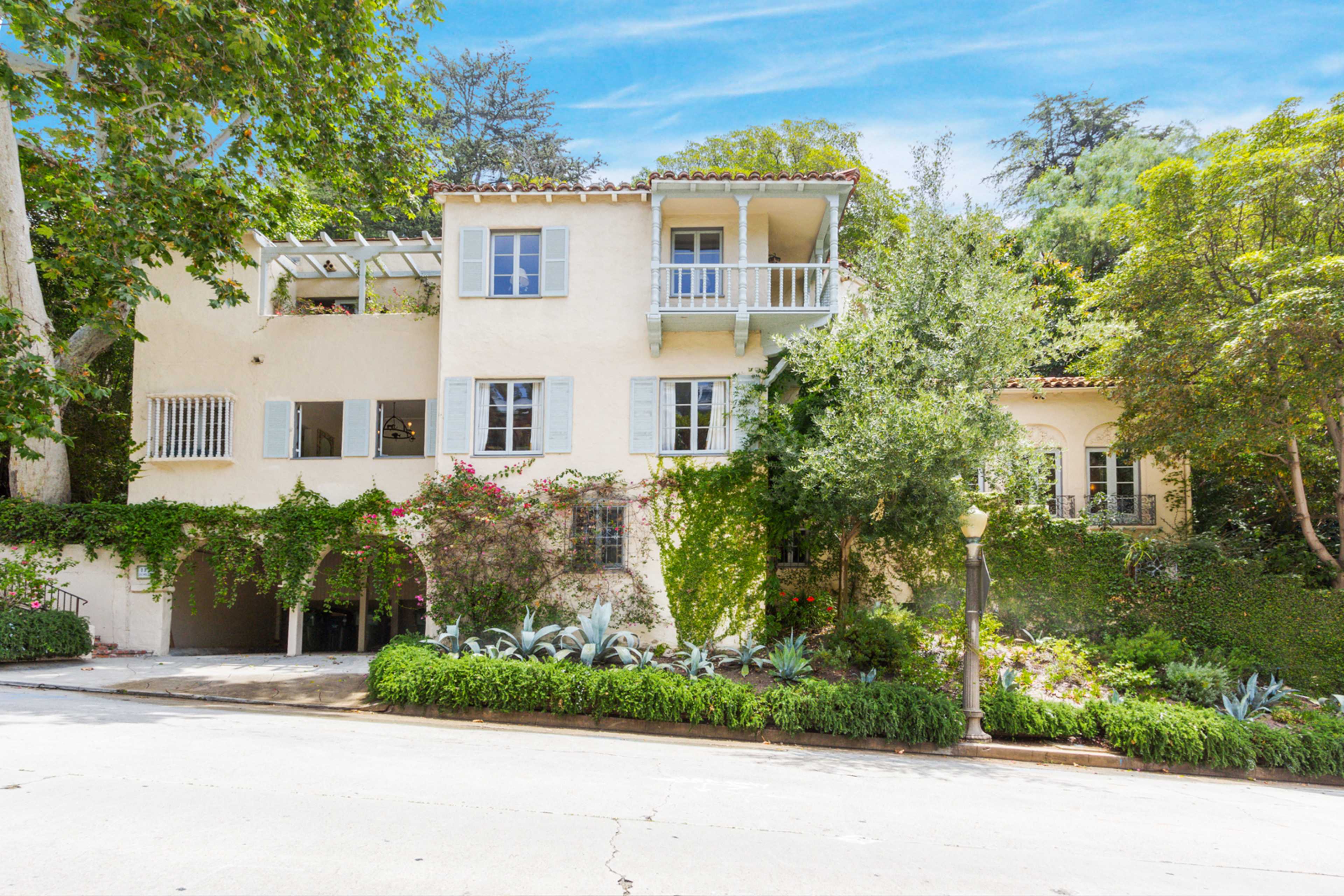 A two-story house with a Mediterranean design features balconies, greenery climbing its walls, and is surrounded by landscaped gardens on a quiet street.