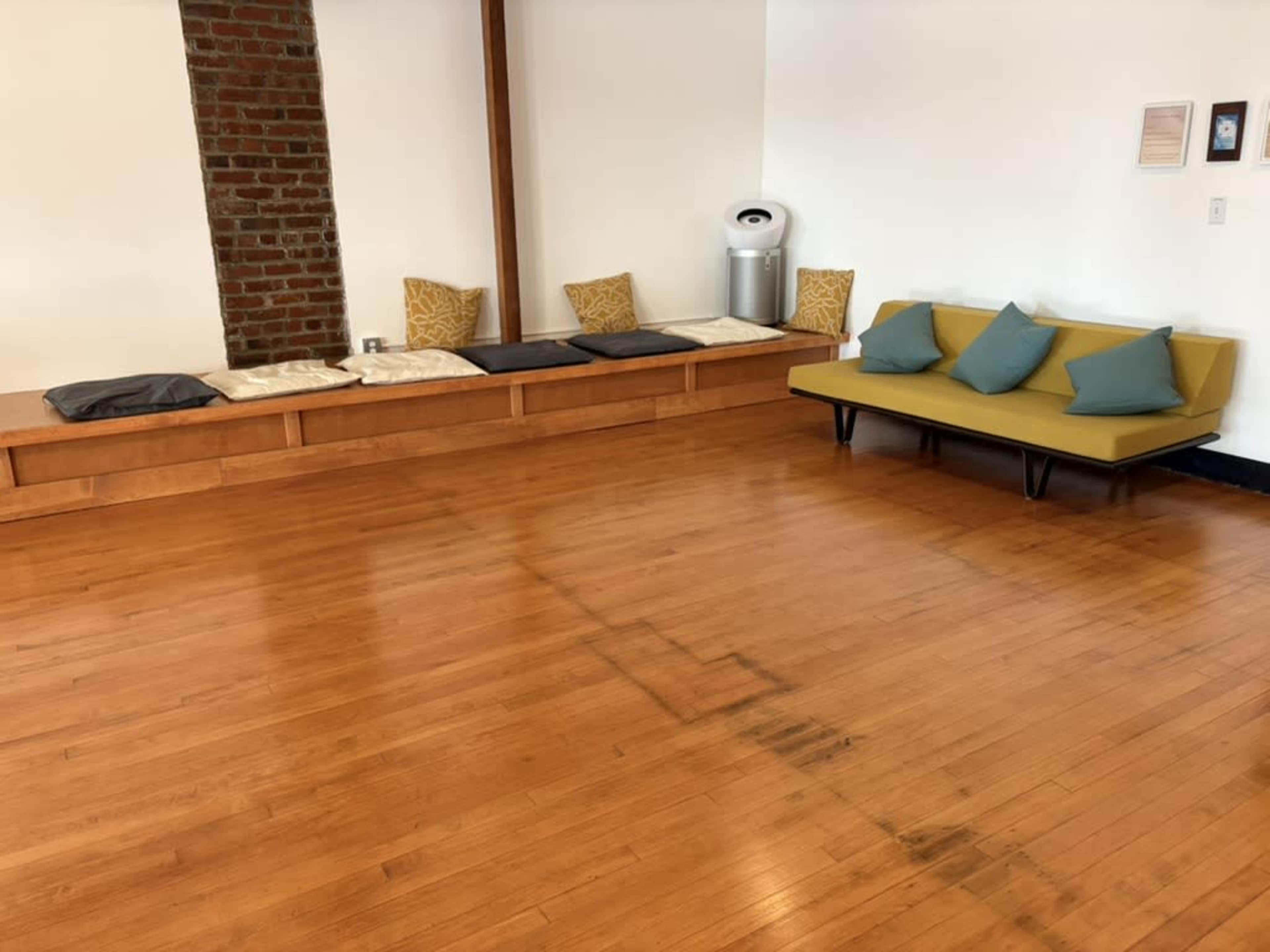 The image shows a spacious room with wooden flooring, a low wooden bench with cushions on one side, and a yellow sofa with blue cushions facing the wall.