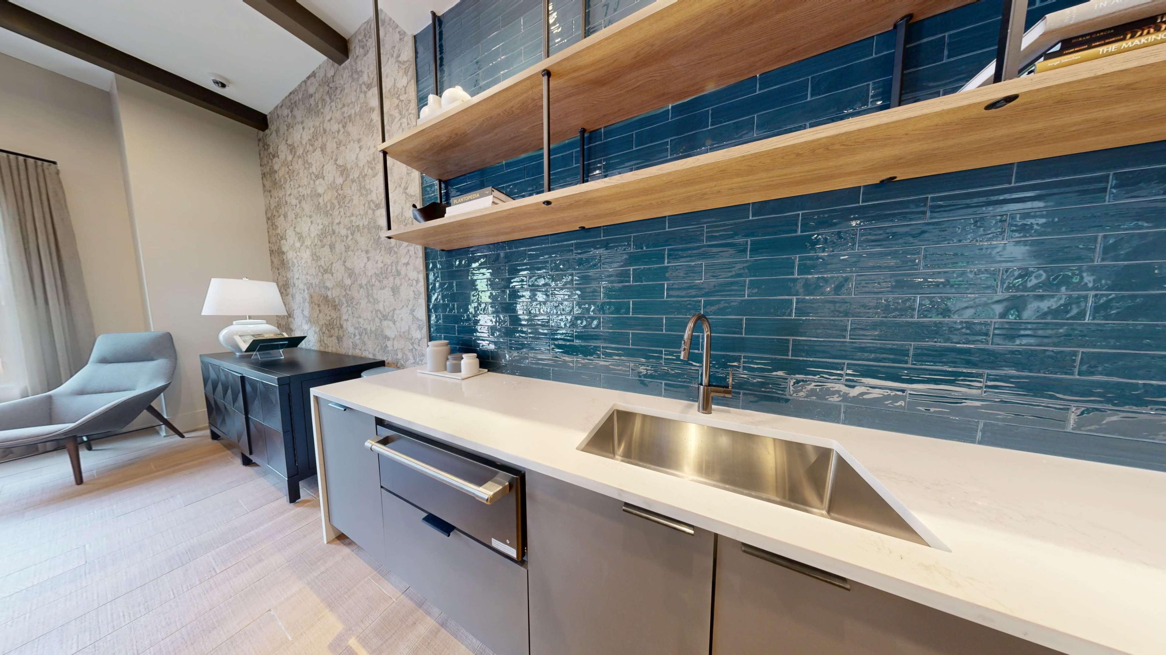 The image shows a modern kitchen setup featuring a stainless steel sink, gray cabinetry, open wooden shelving, and a blue tiled backsplash.