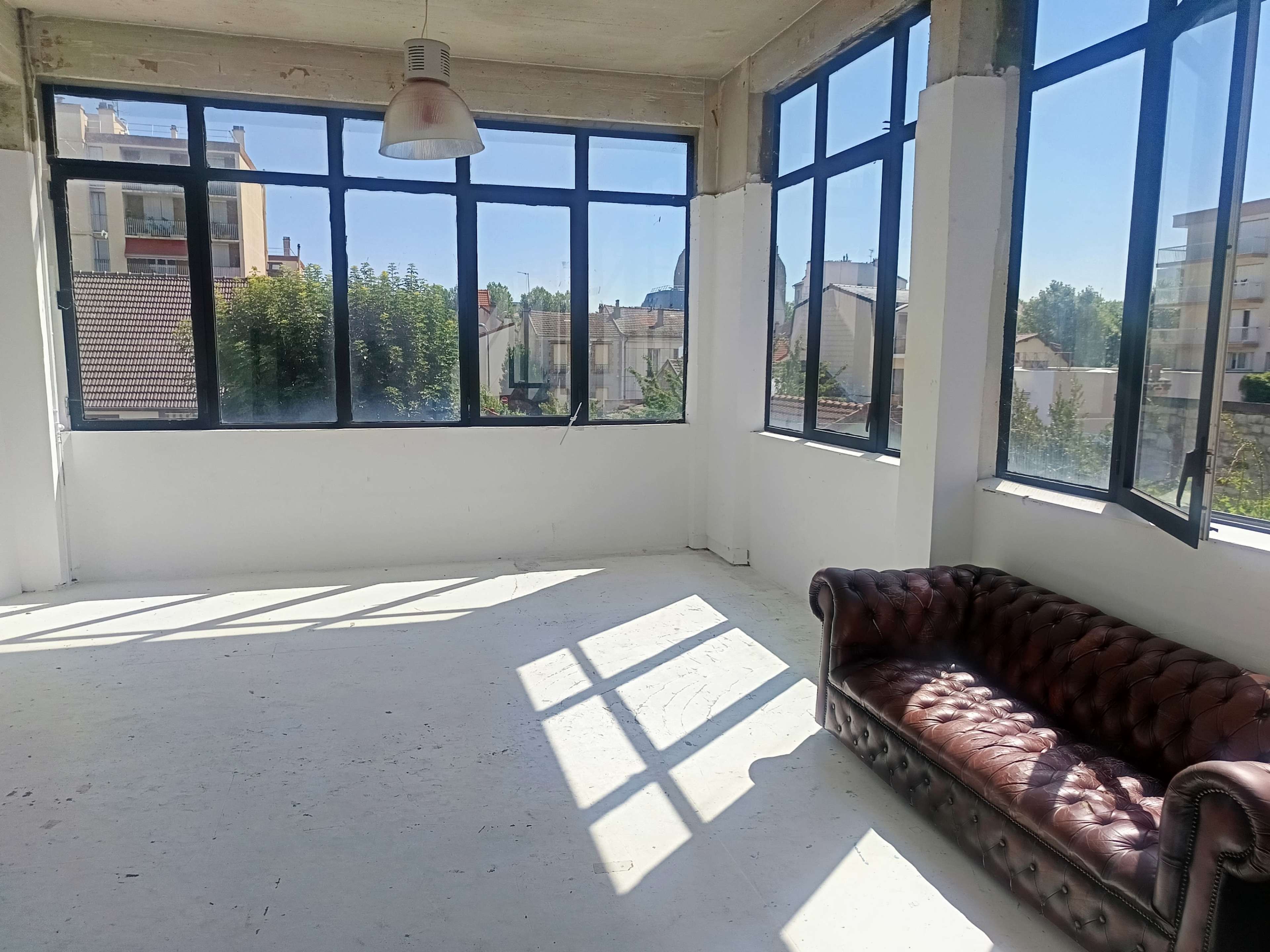 The image shows a bright, empty room with large windows letting in natural light and a brown leather sofa.
