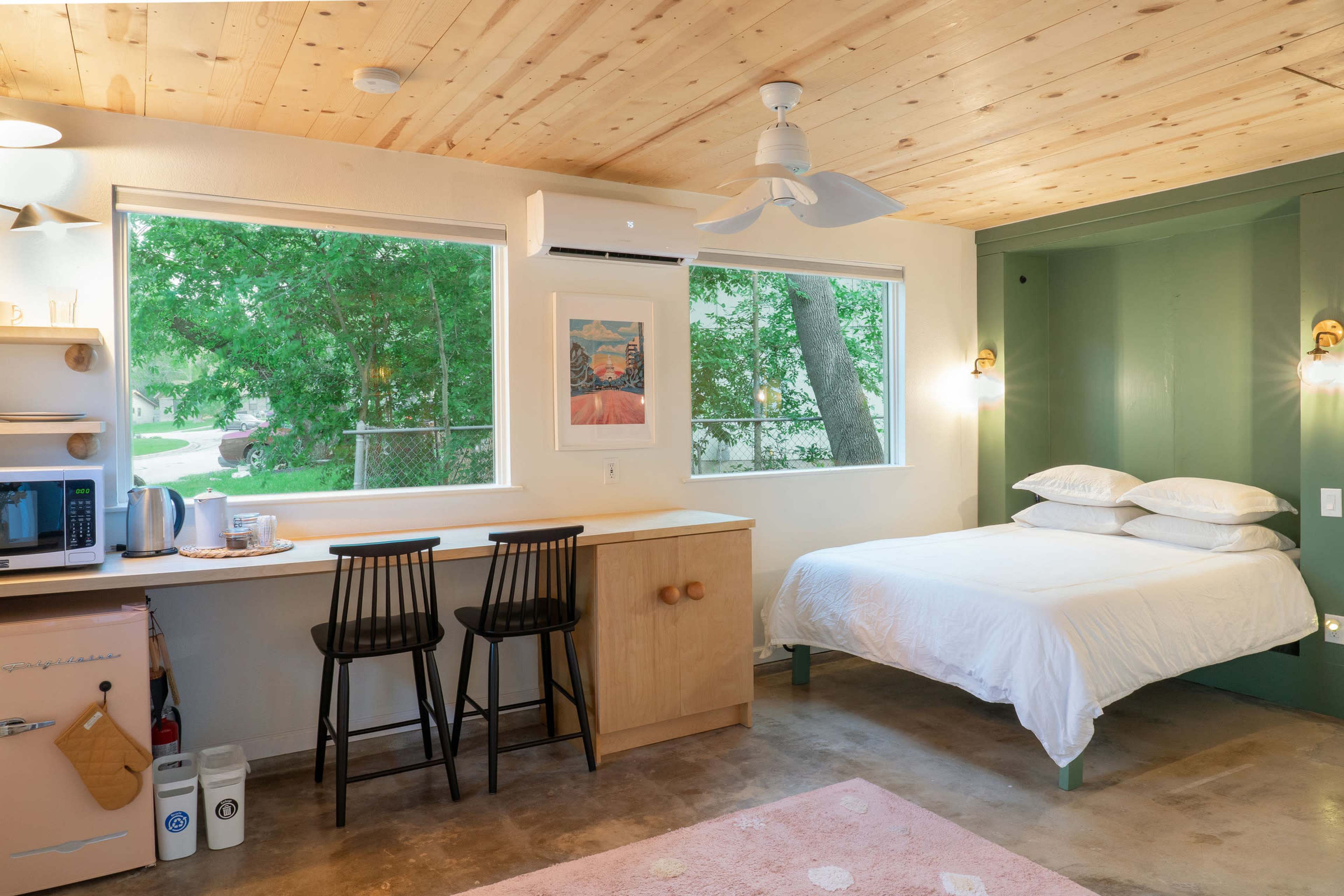 A modern, minimalistic bedroom with a bed, a small kitchenette with two bar stools, and large windows allowing natural light to enter.