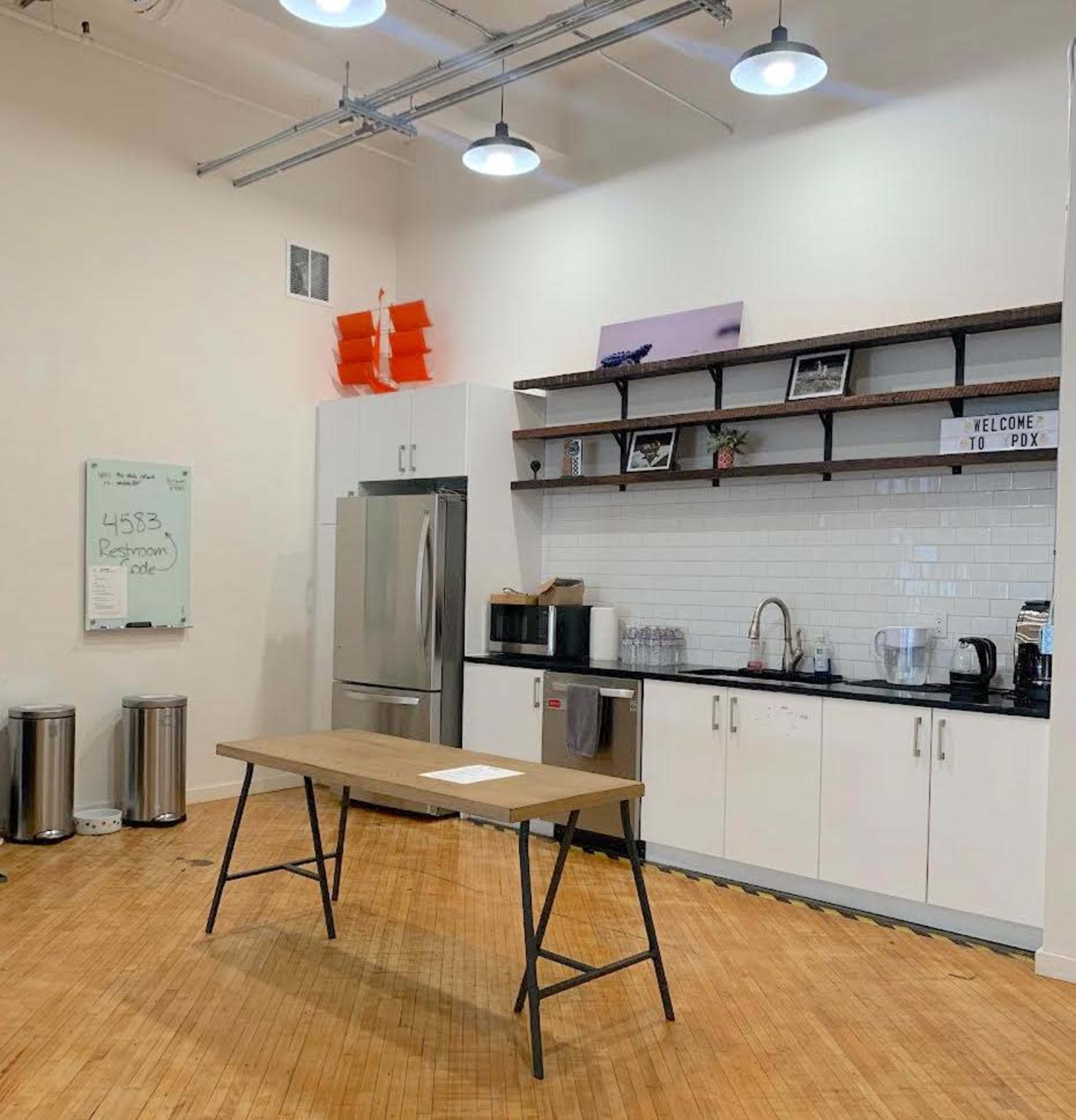 A modern kitchen space with a table in the center, a stainless steel refrigerator, a sink, and shelves displaying various kitchen items.