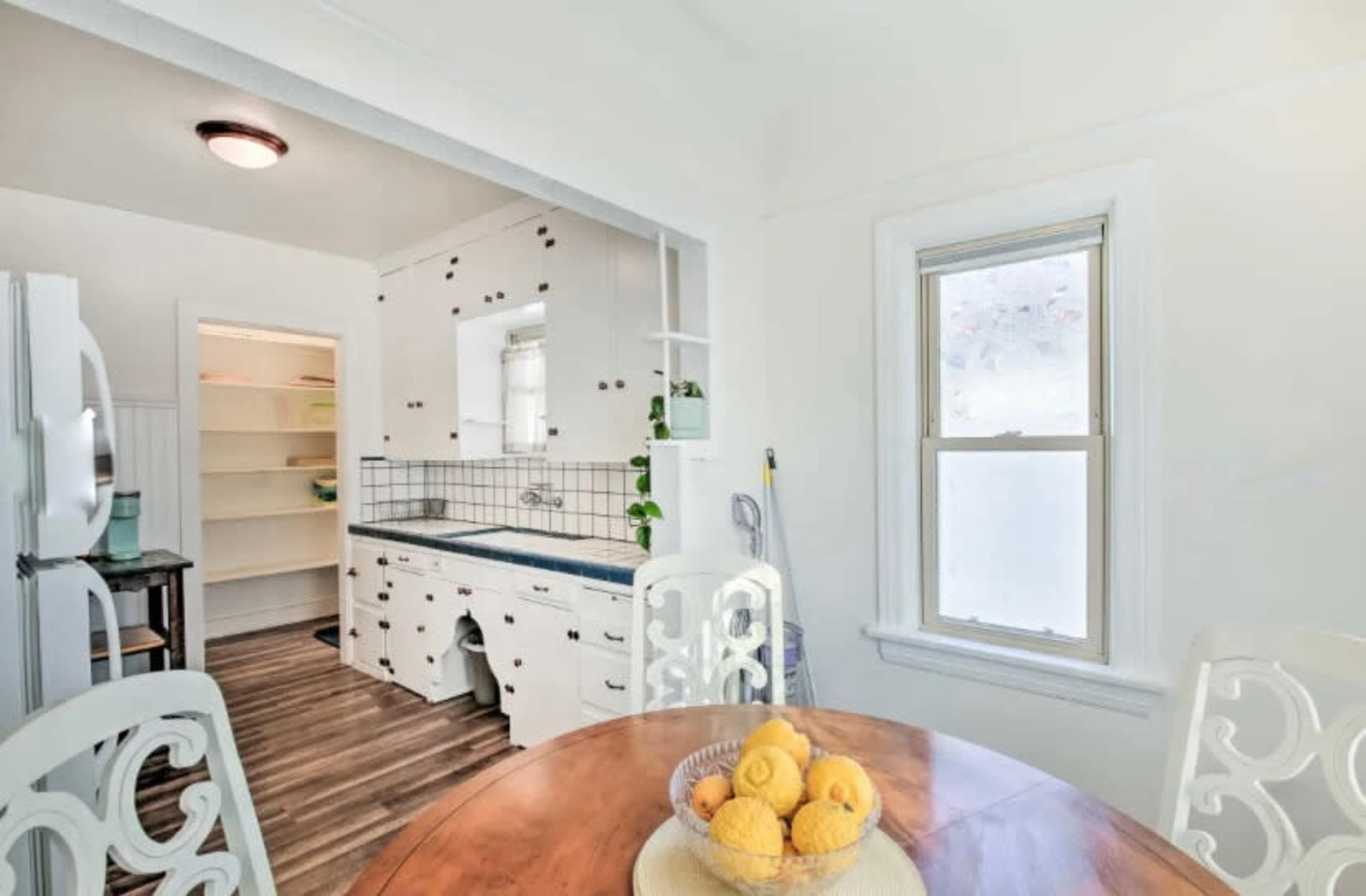 A bright kitchen features a wooden table with a bowl of lemons, surrounded by white chairs, and includes an adjoining pantry area.