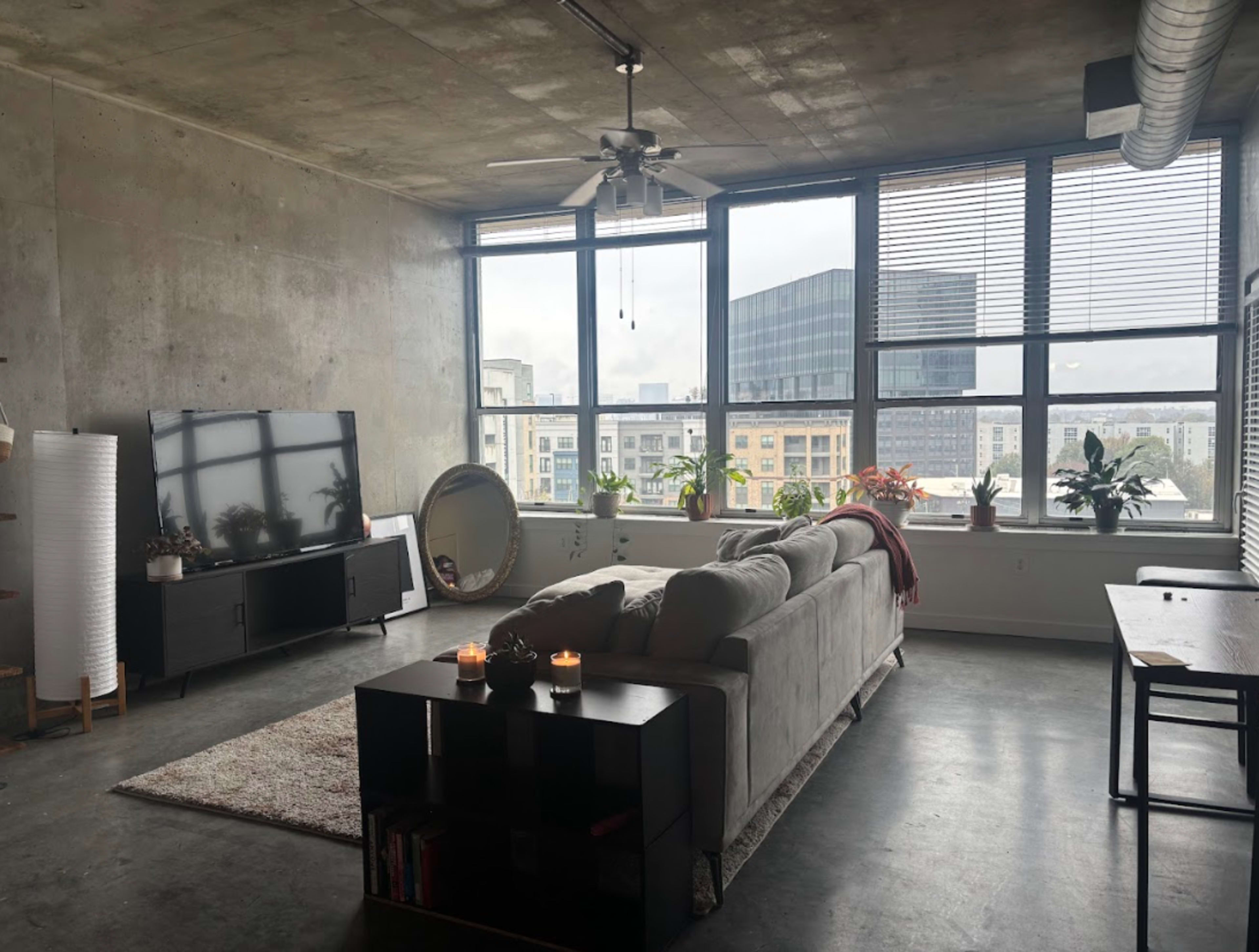 The image shows a modern living room with a gray sectional sofa, a television on a black stand, and large windows offering a view of a cityscape.
