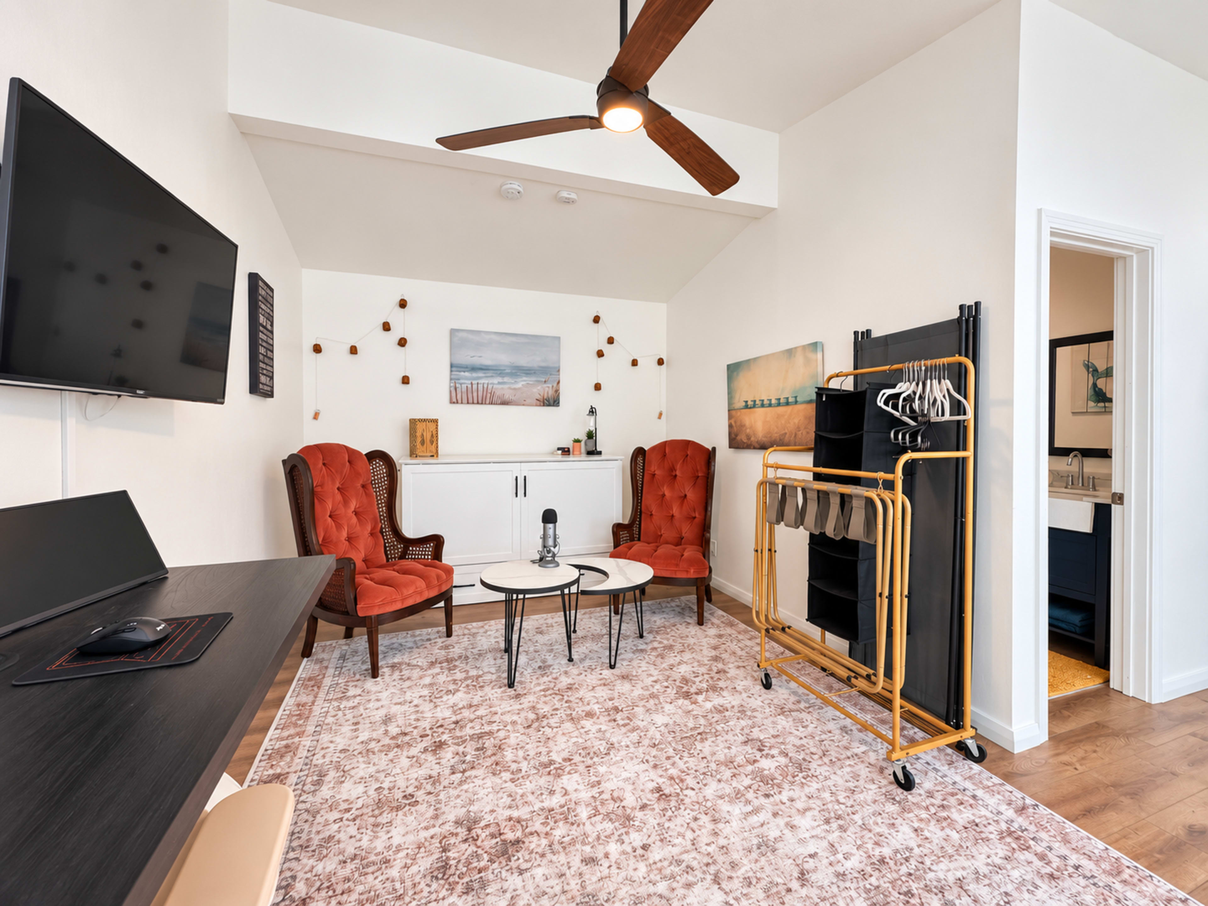 The room features two red armchairs, a small coffee table, a wall-mounted television, and a gold rolling rack with hanging space, complemented by a textured area rug.