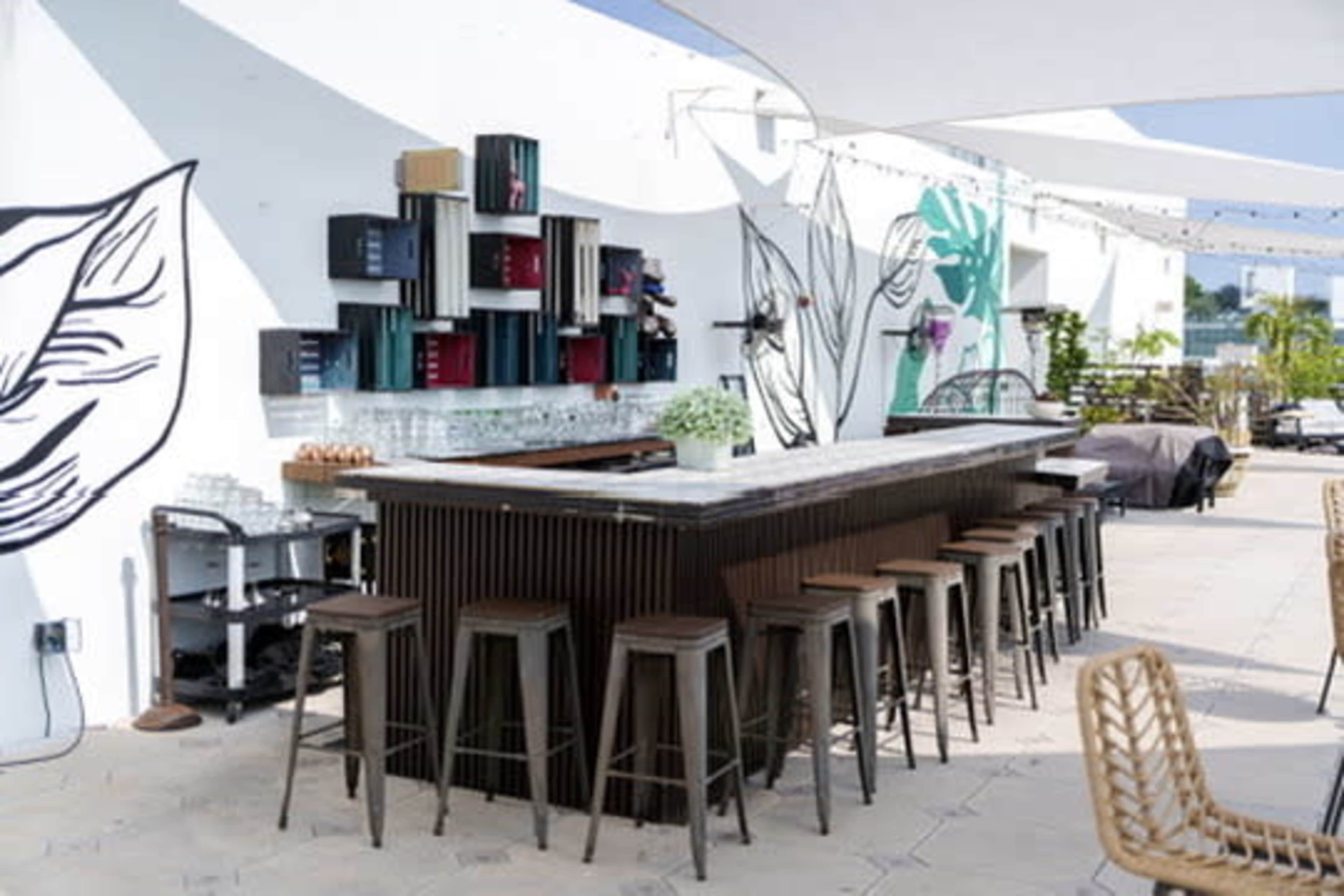 The image shows a modern outdoor bar with a marble countertop, surrounded by tall metal stools, against a backdrop of colorful wall murals.