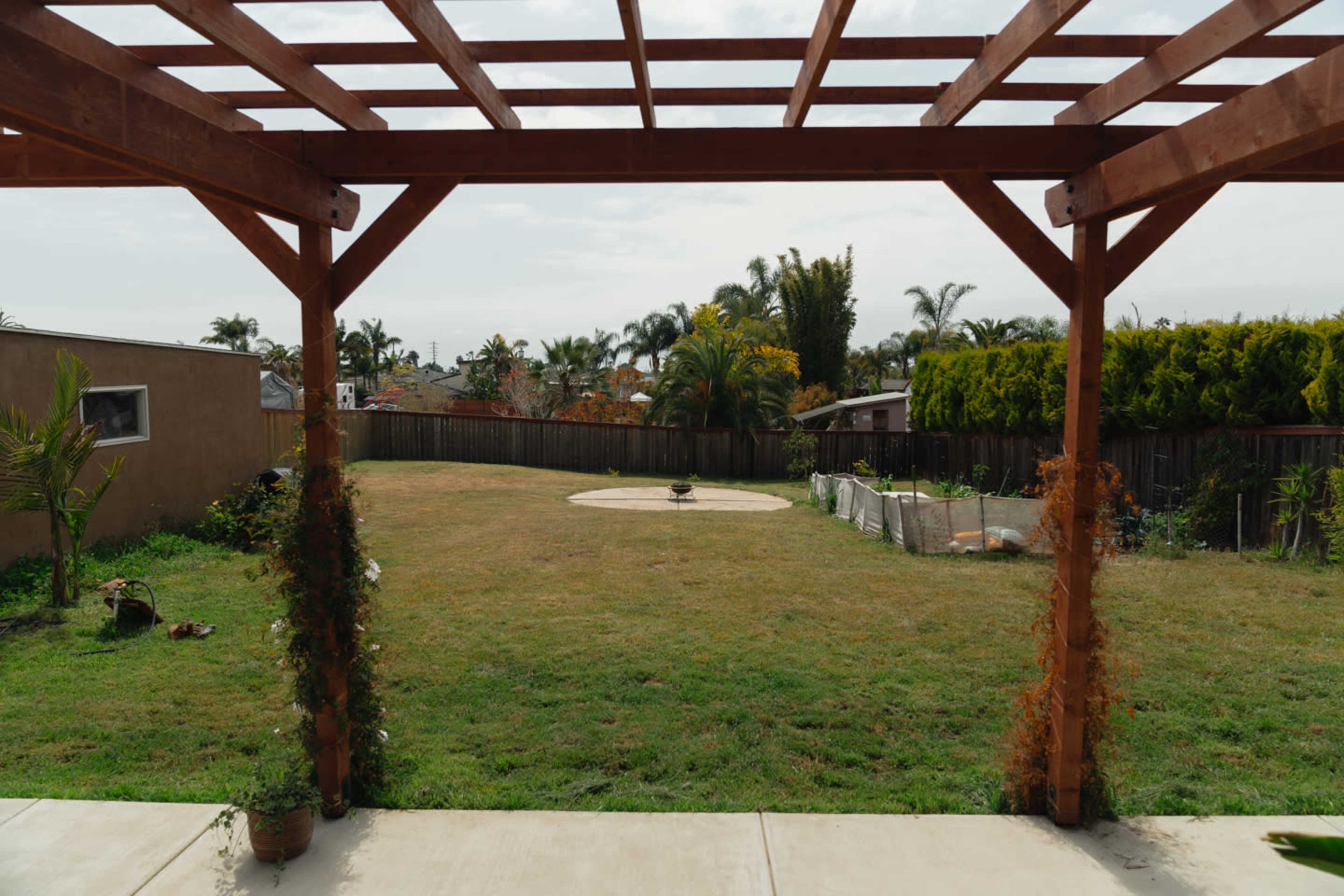 A backyard patio framed by a wooden pergola overlooks an expansive grassy area with a circular stone feature in the center.