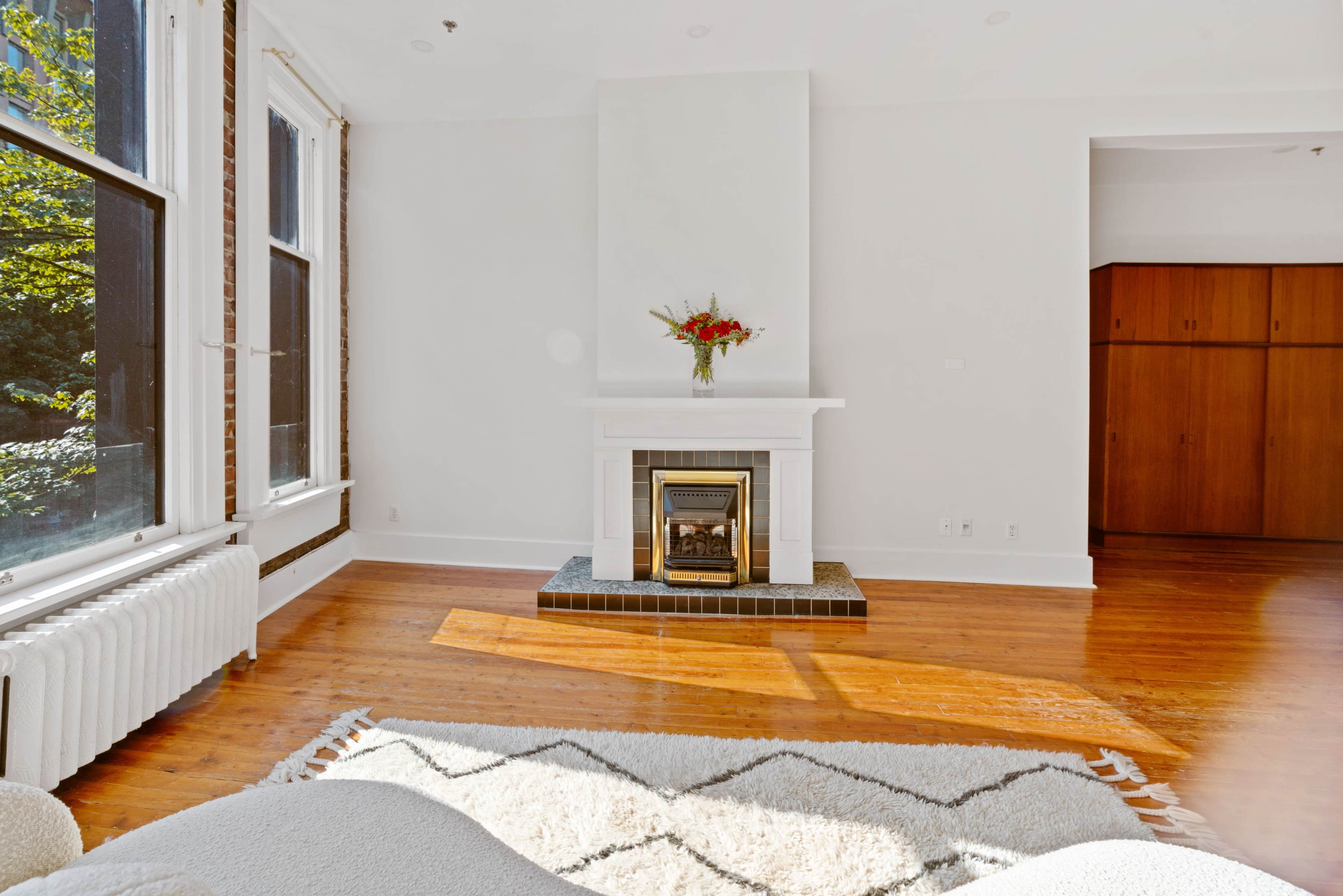 A bright living room features a white fireplace with a vase of flowers and large windows letting in natural light, next to a wooden closet.