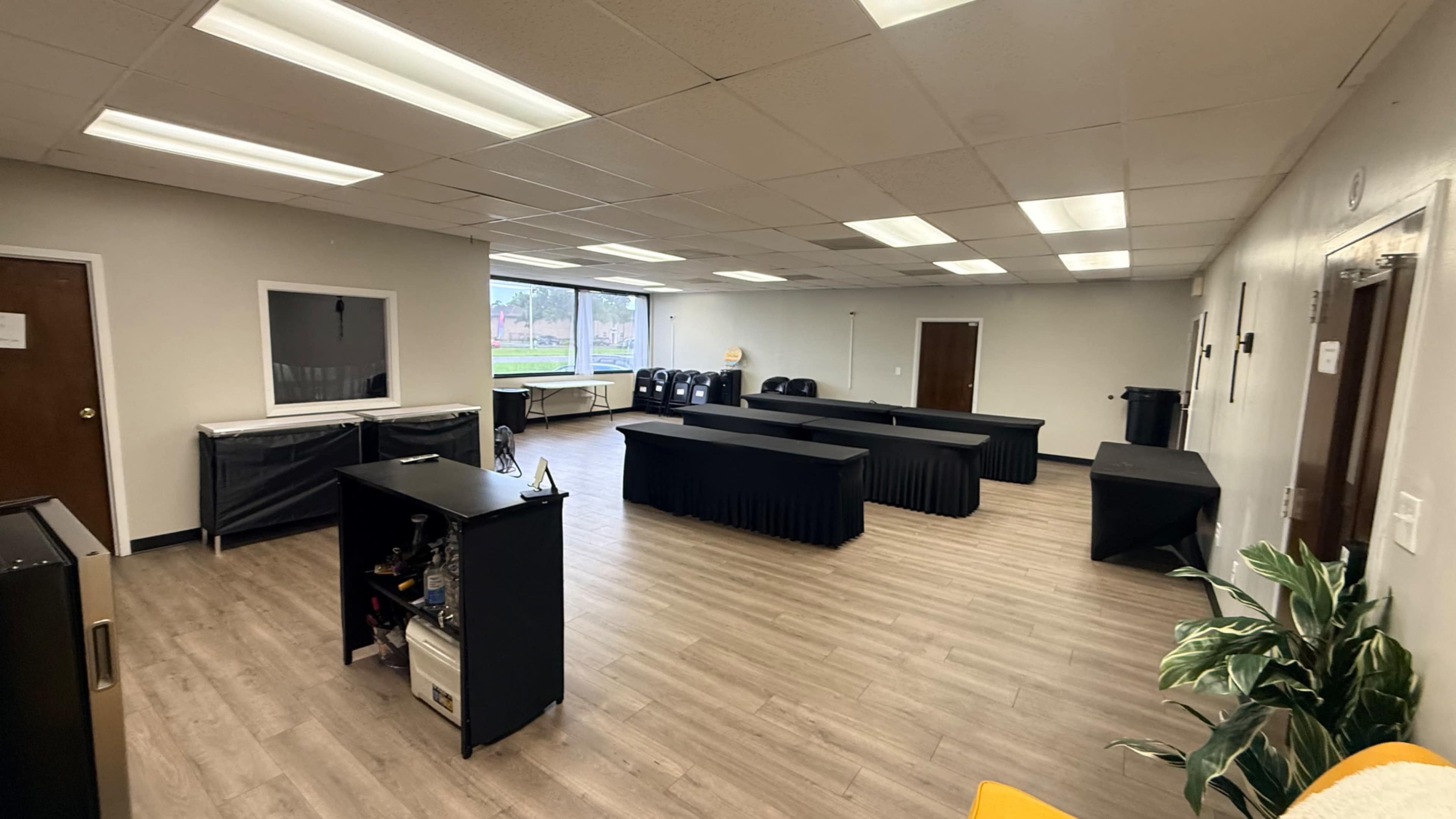 The image shows a spacious room with wooden flooring, several tables covered in black cloth, and large windows allowing natural light to enter.