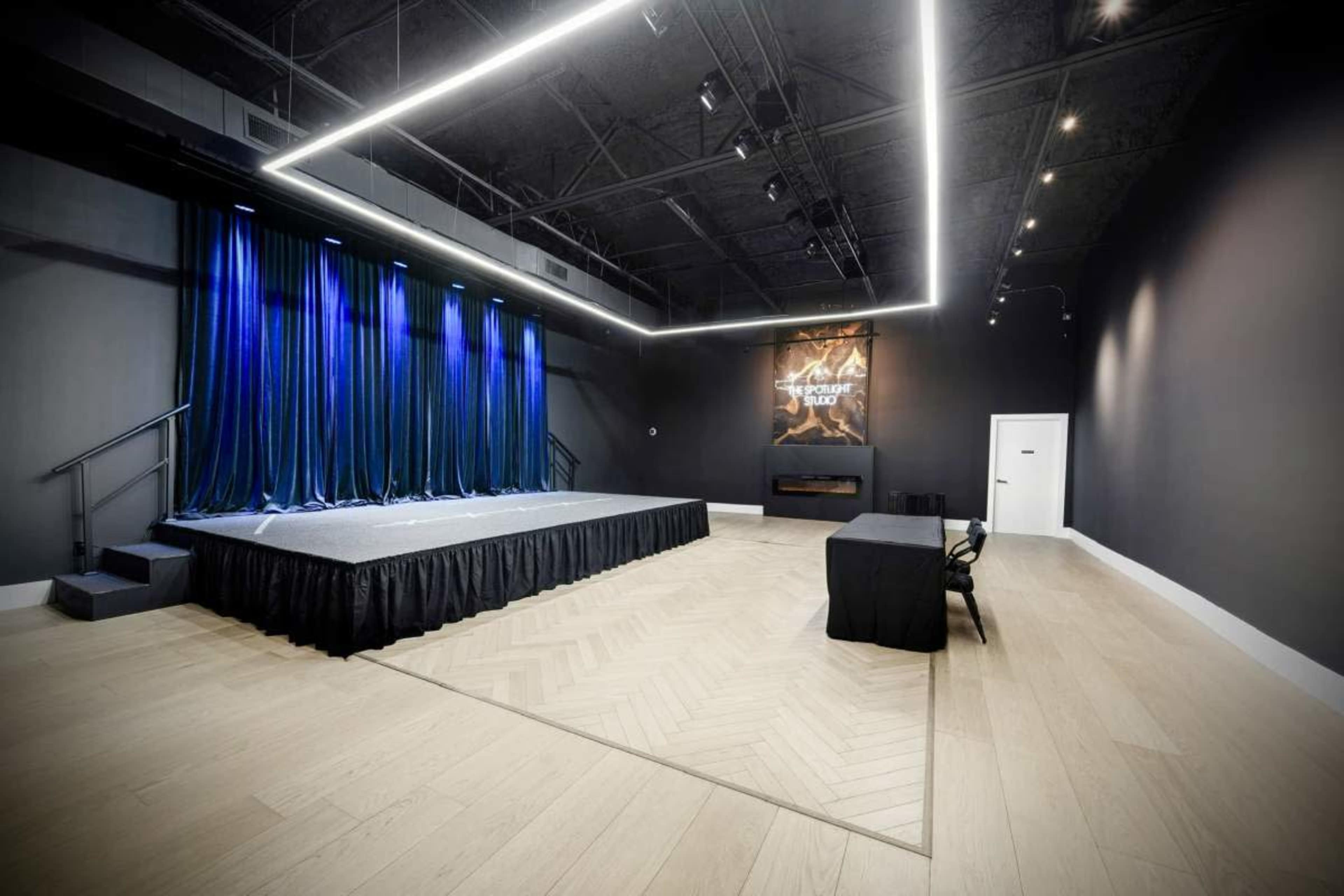 The image depicts a spacious performance stage with dark blue curtains and a simple black table and chair set against a gray wall.