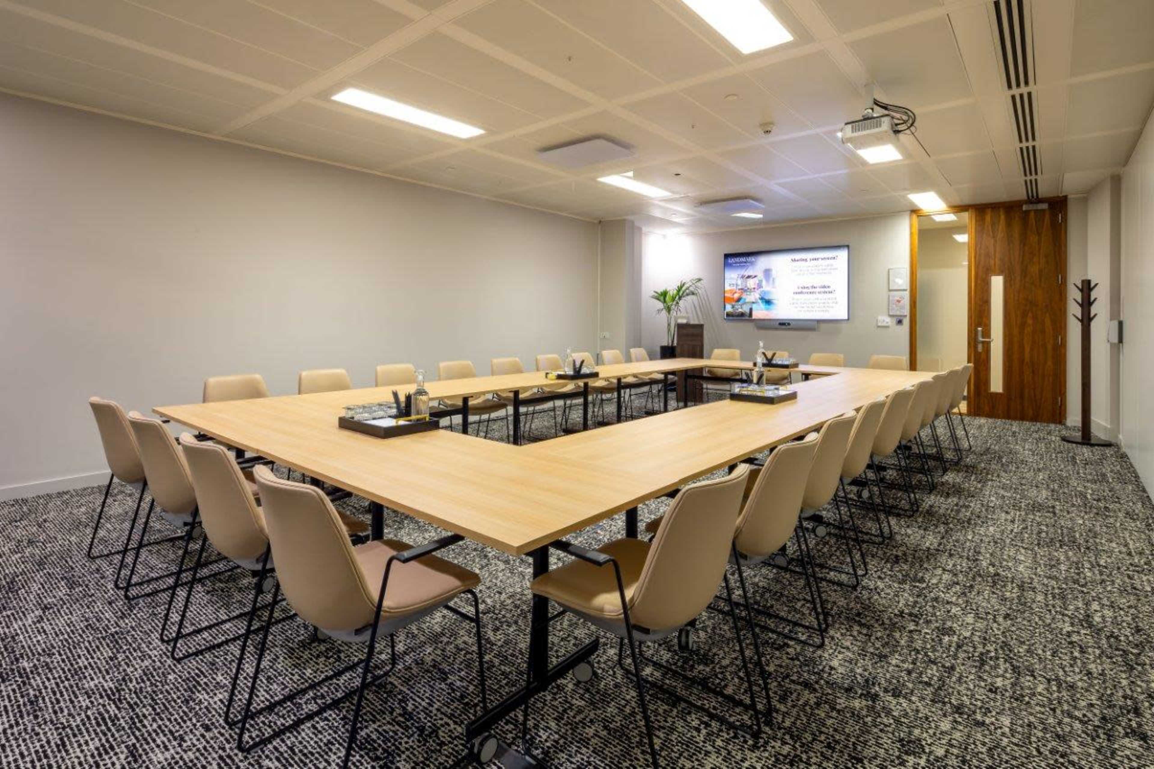 A rectangular conference room setup with a long table surrounded by chairs, a presentation screen at the front, and a plant in the corner.