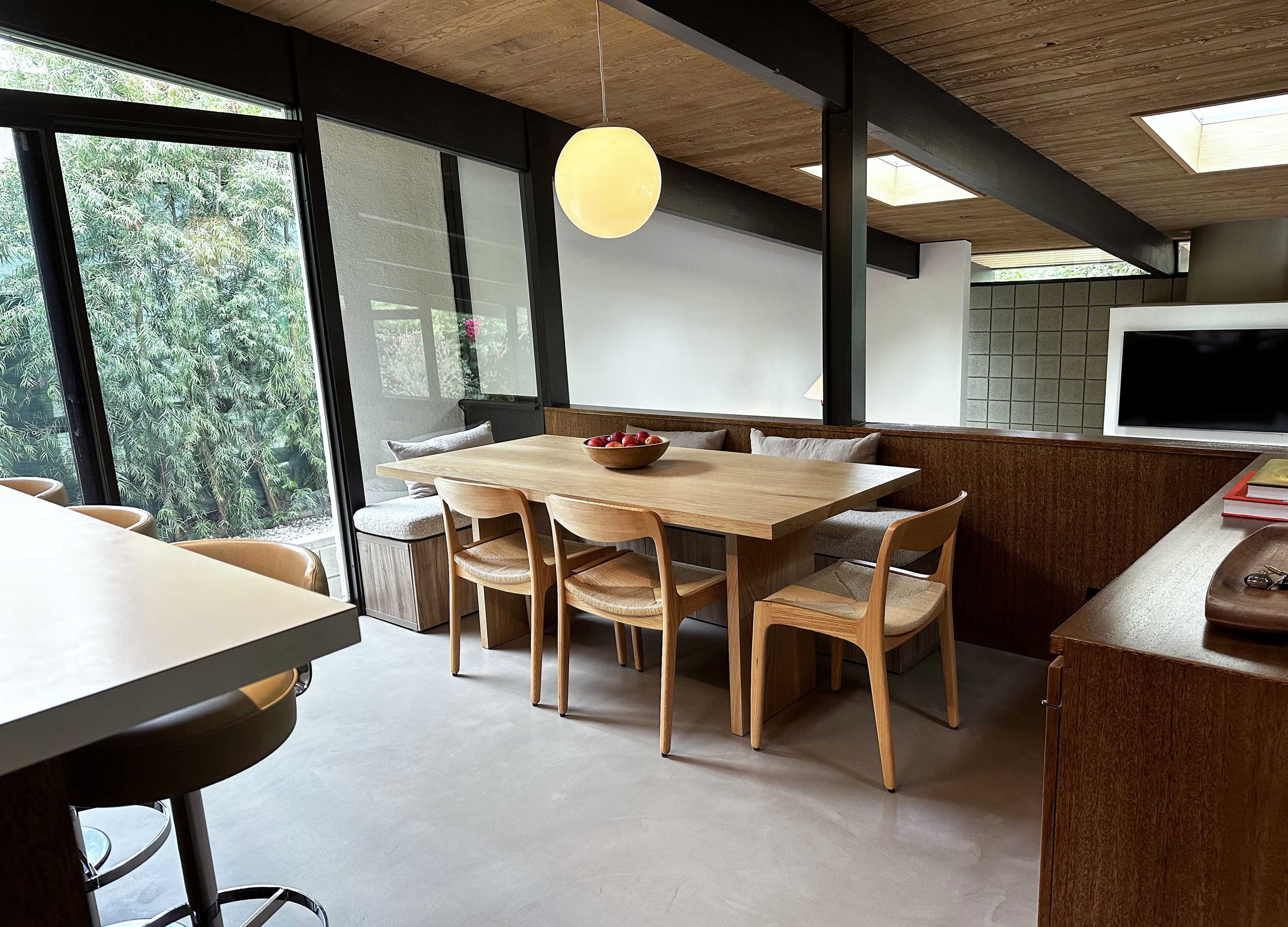 A modern dining area features a wooden table surrounded by chairs, with large windows providing views of greenery outside.