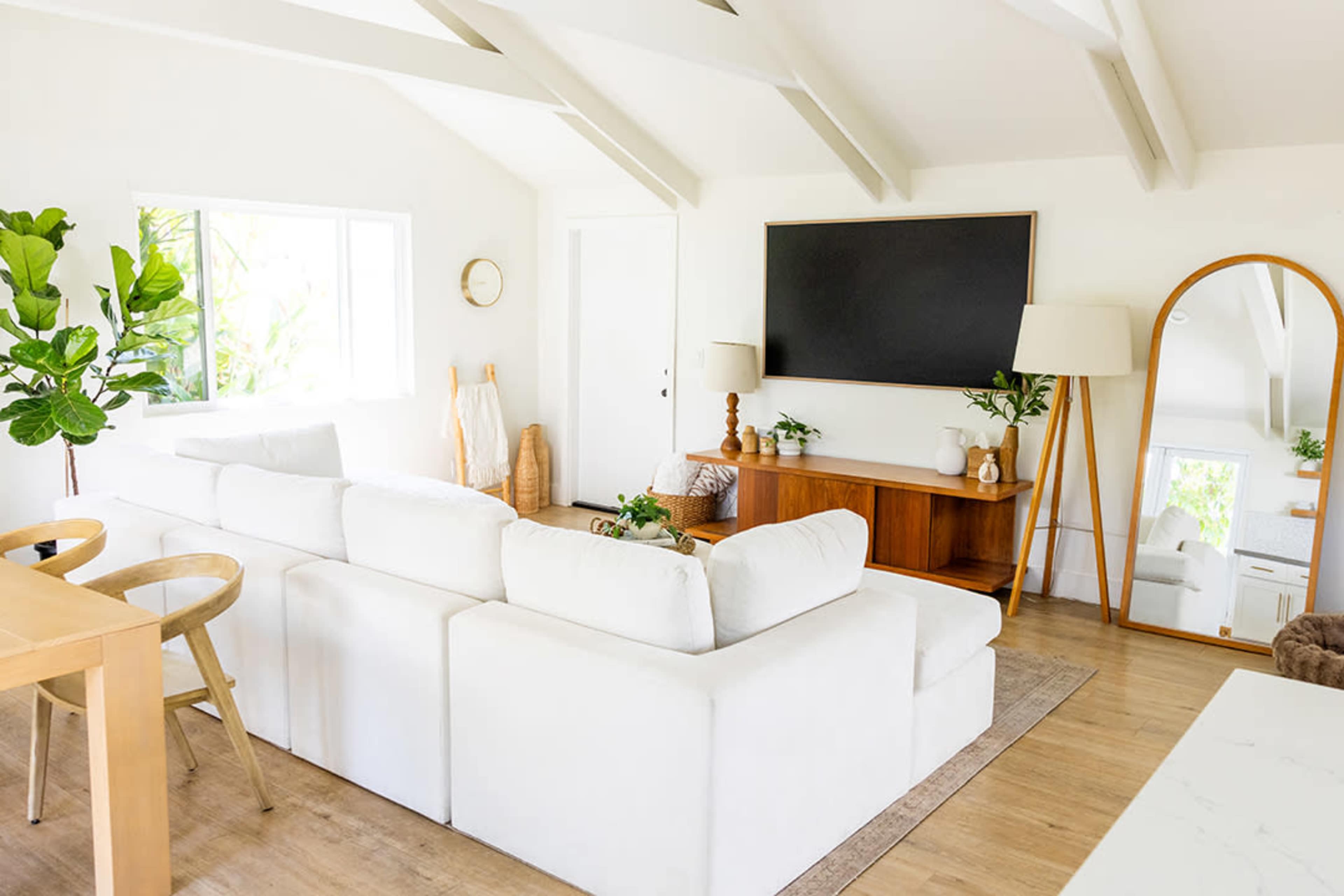 The image shows a bright, minimalist living room featuring a white sectional sofa, a wooden side table, a large wall-mounted television, and a tall mirror.