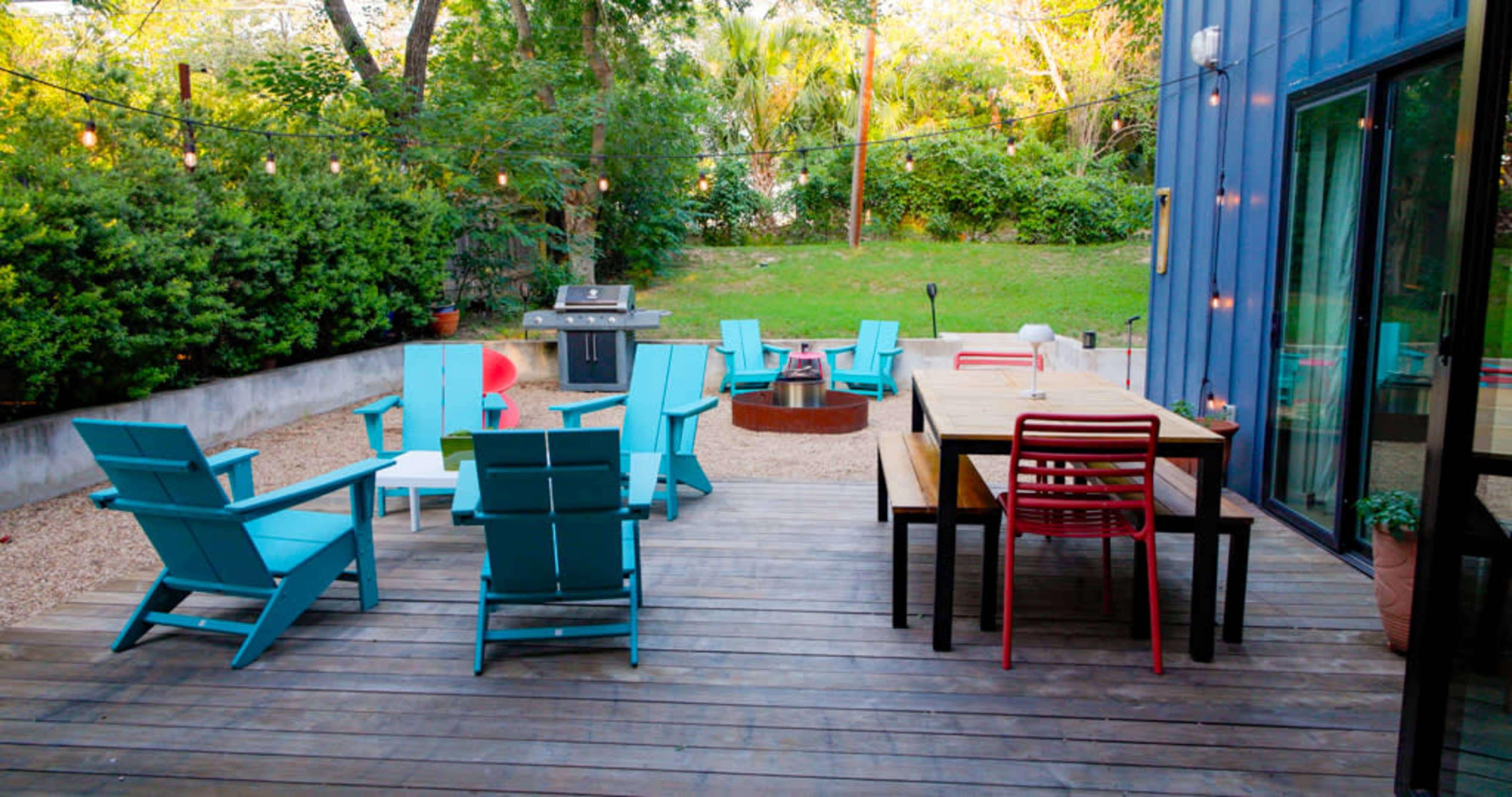 The image shows a wooden deck with several turquoise chairs, a dining table, and a grill, surrounded by greenery and string lights.