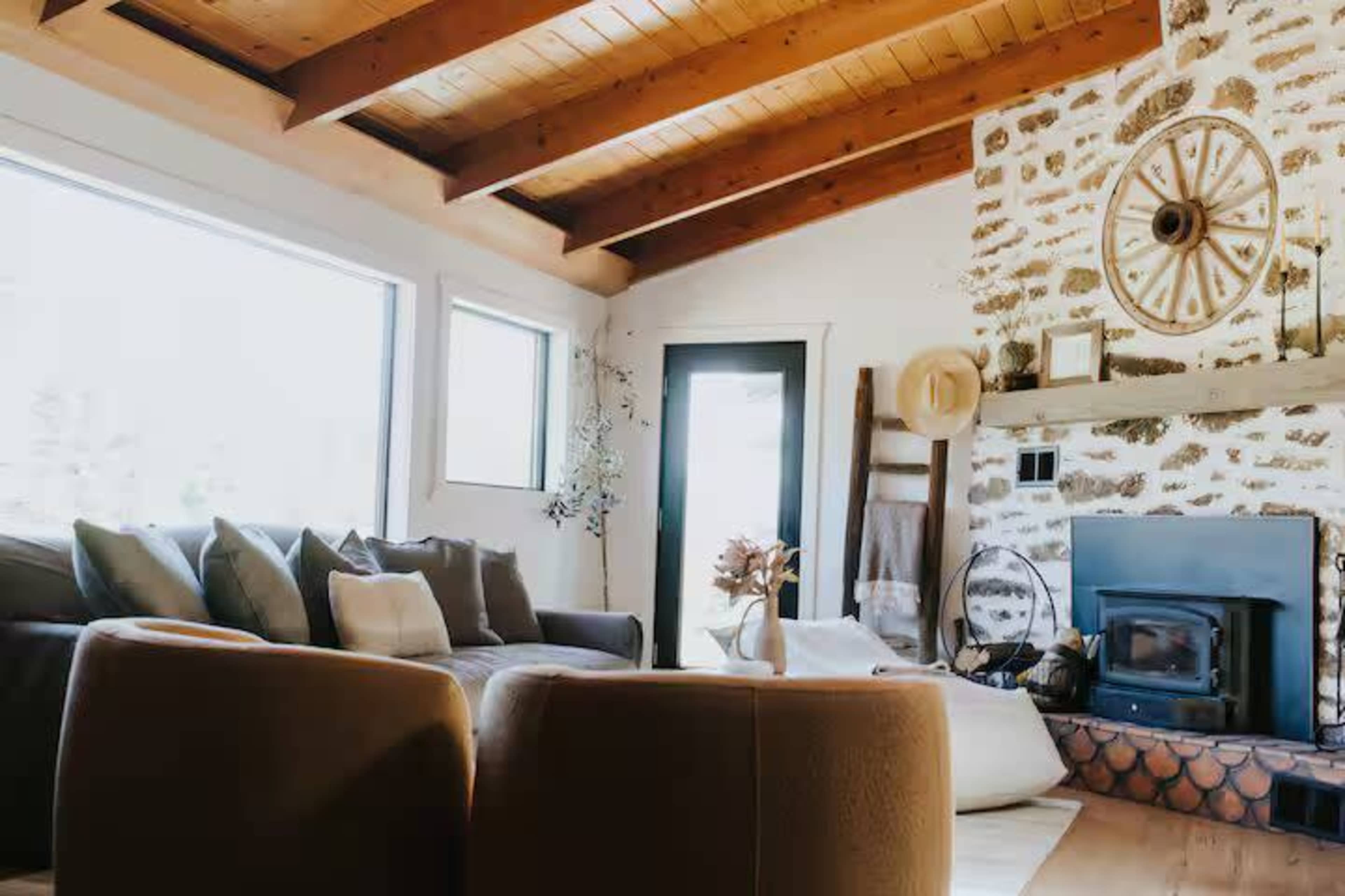 The living room features a stone fireplace, wooden beams, large windows, and a cozy seating arrangement with cushions.