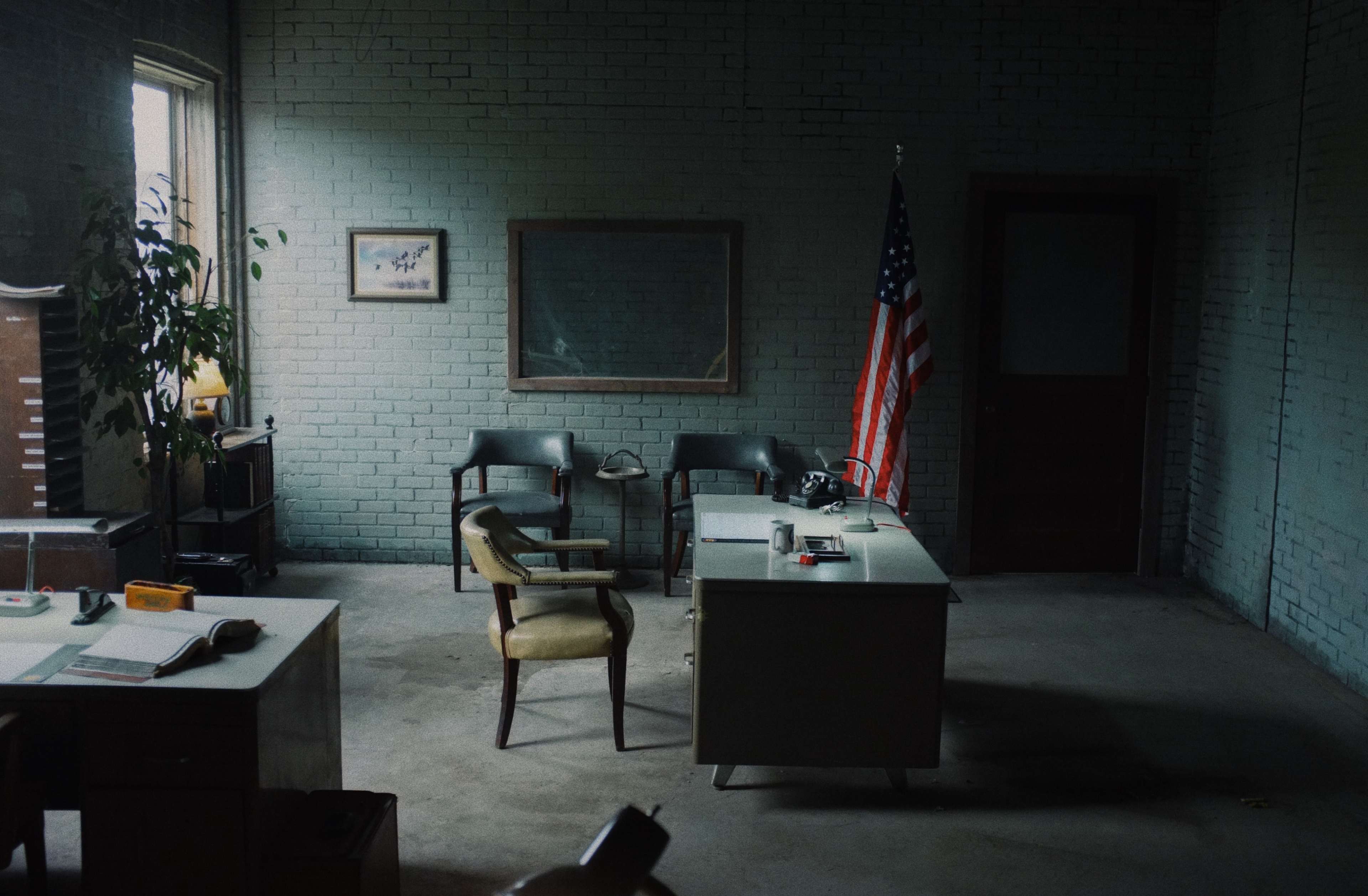 The image shows a sparsely furnished office space with an American flag, two chairs, a desk, and a window letting in natural light.