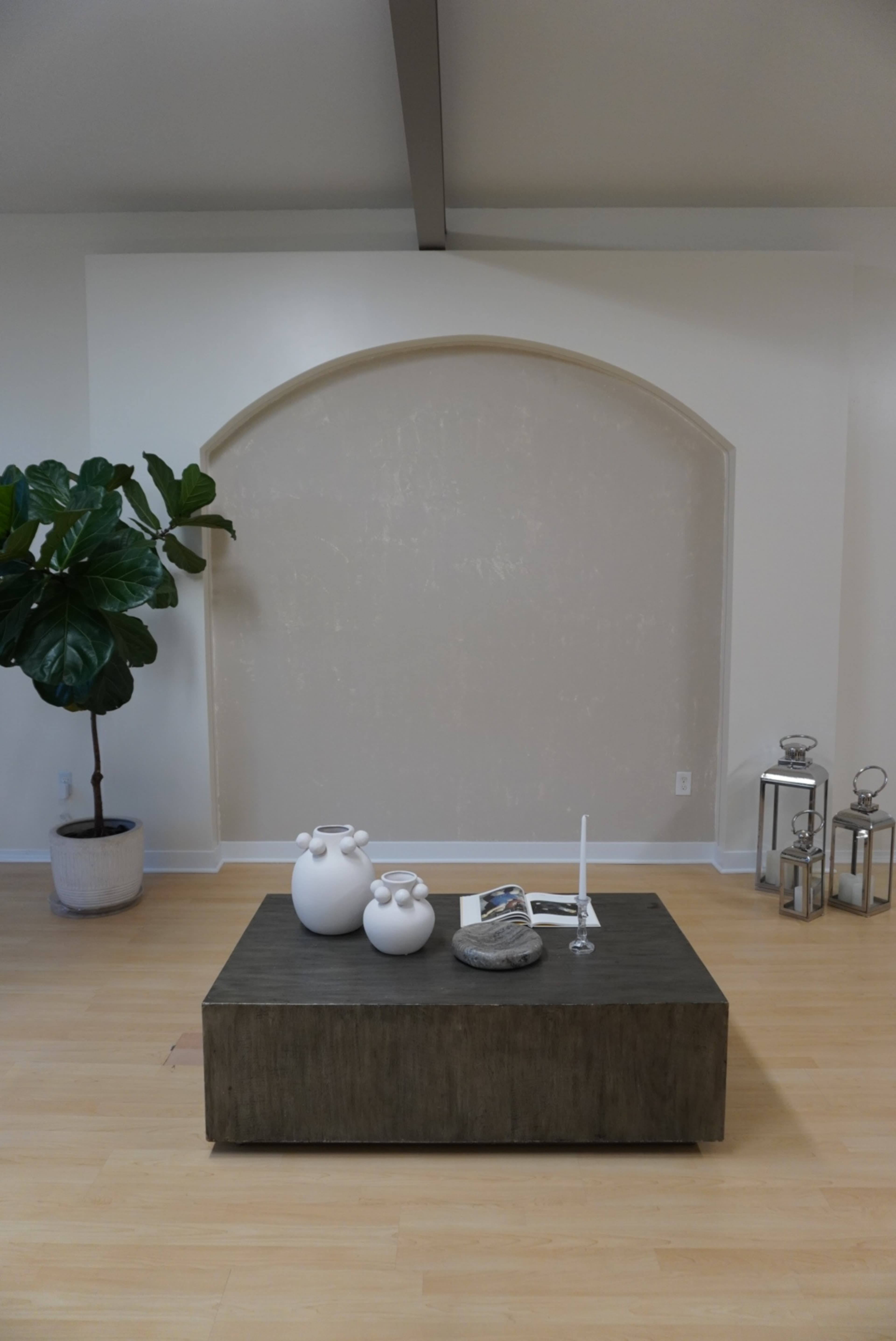 The image shows a minimalistic living room with a wooden coffee table in the center, two decorative vases on top, and a large indoor plant beside an arched wall.