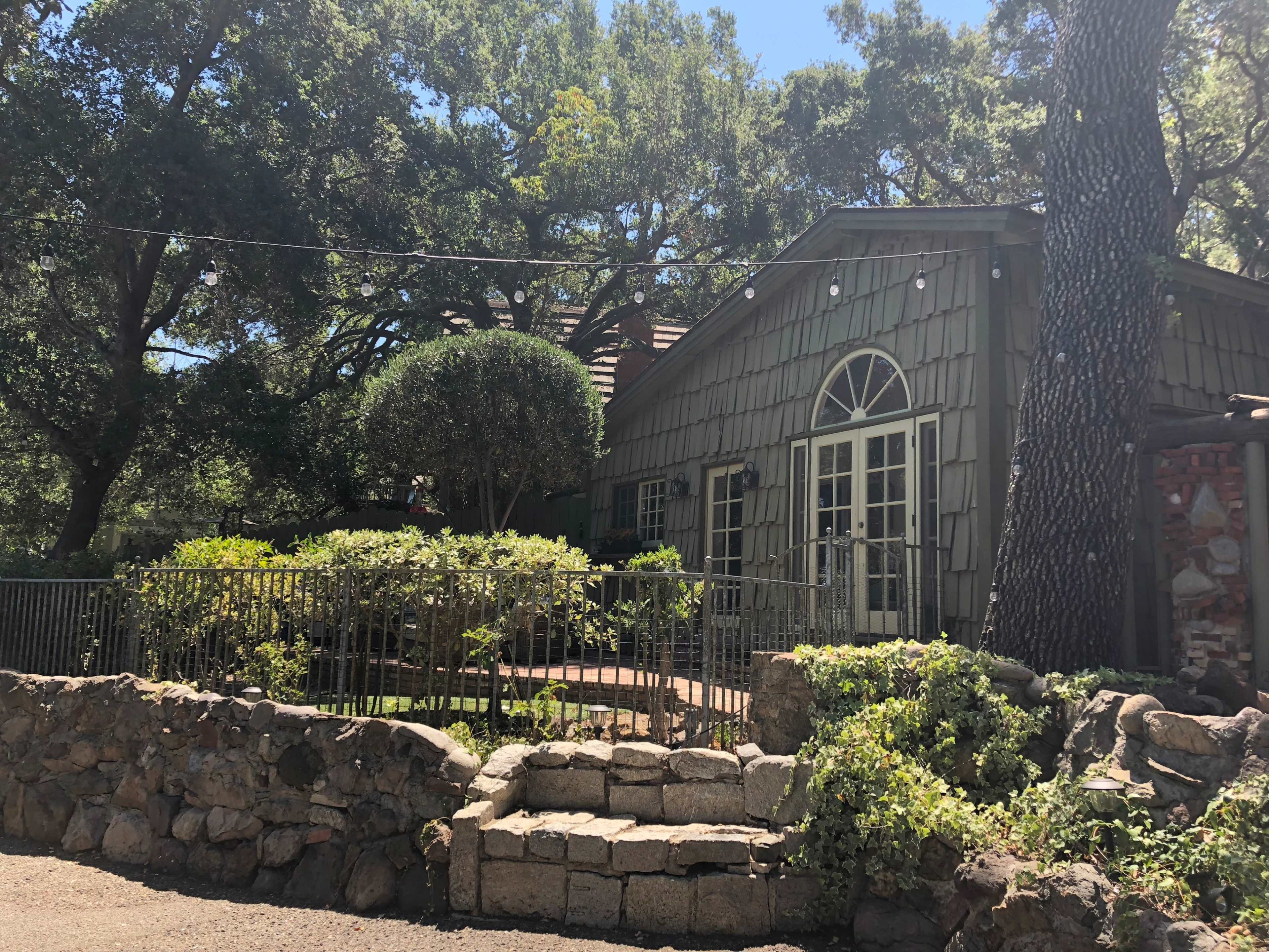 A green, wooden house with large windows is surrounded by trees and a neatly trimmed garden, featuring a stone wall and pathway.