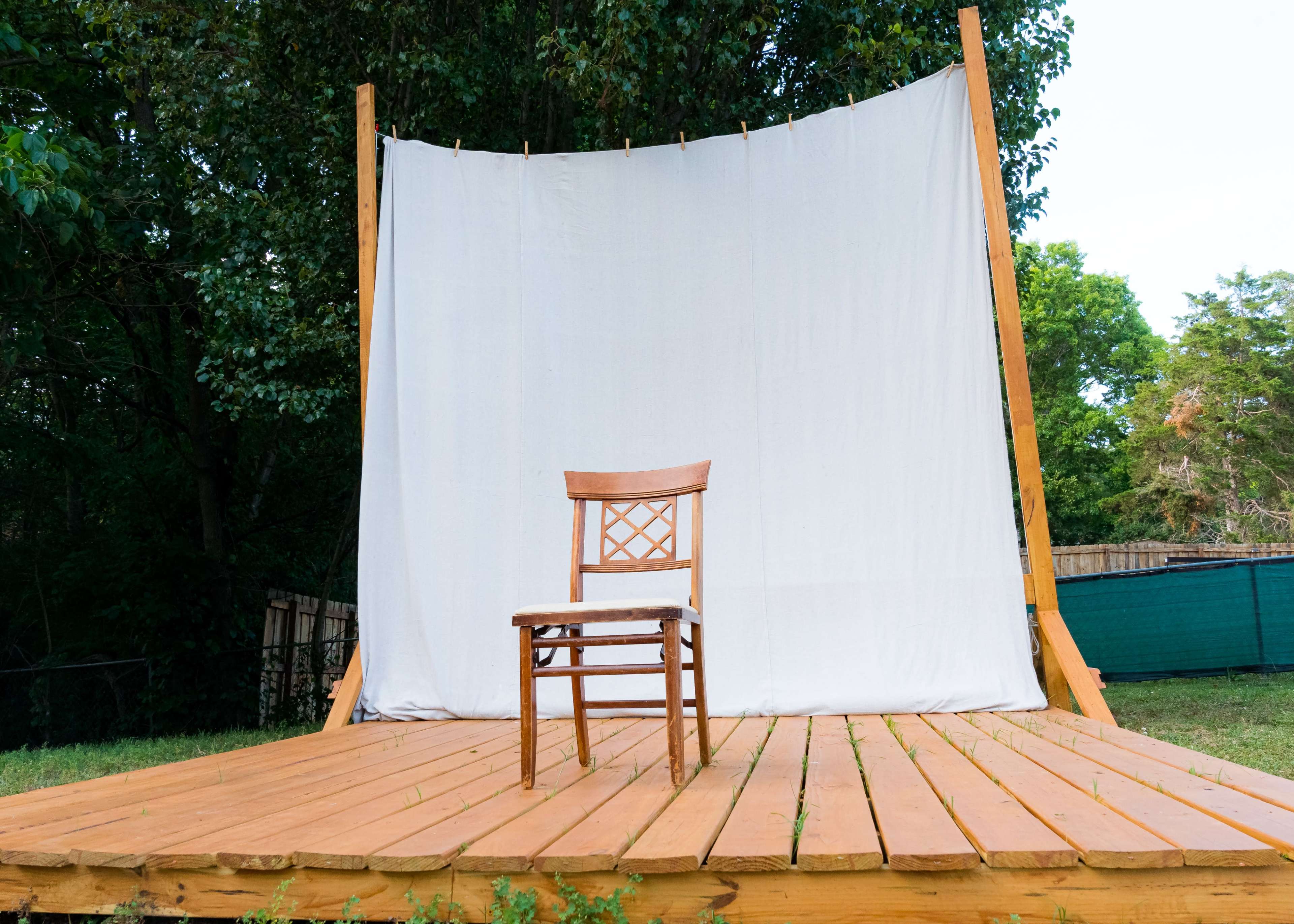 A wooden chair stands alone on a wooden stage framed by a white backdrop and surrounded by greenery.
