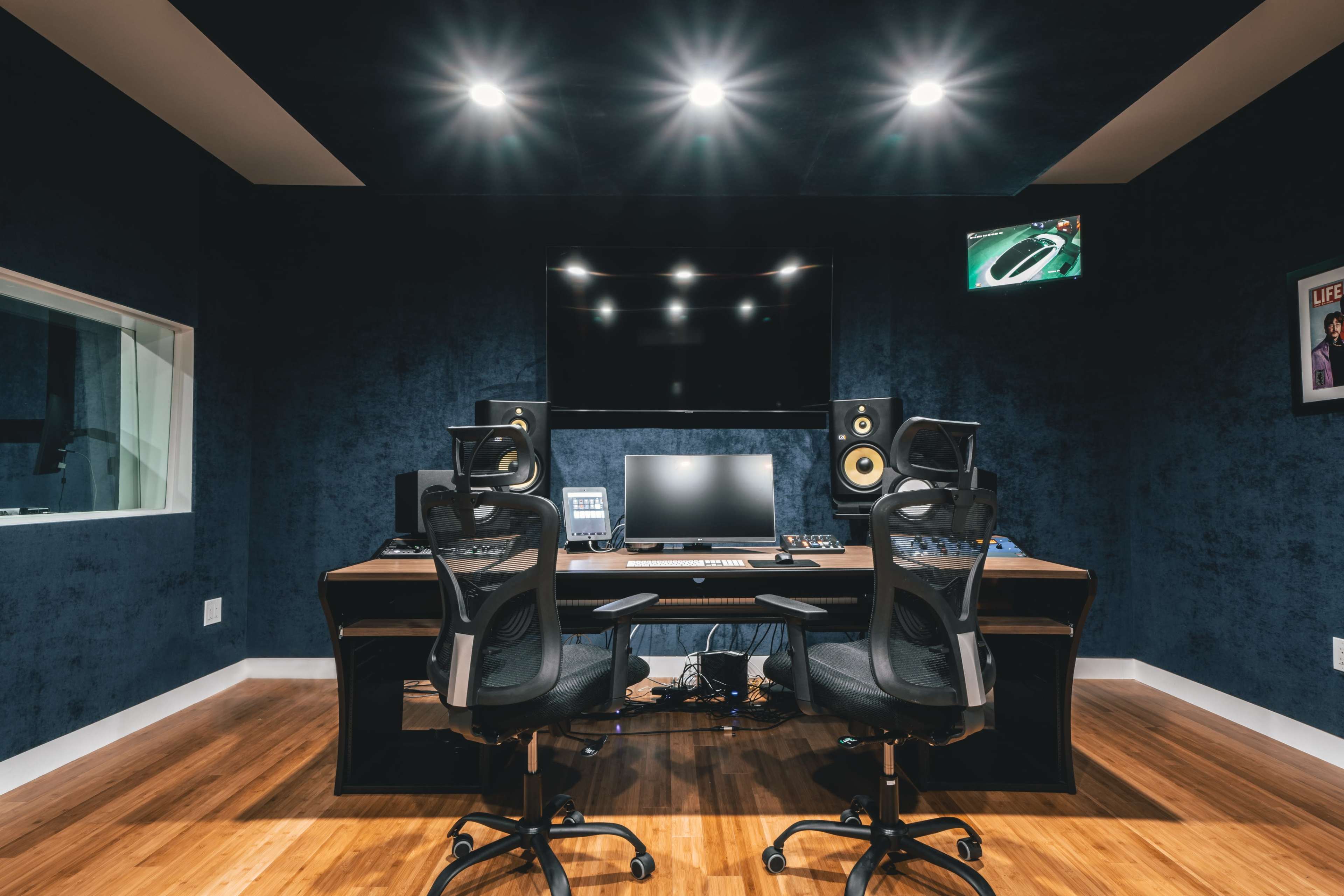 A modern recording studio equipped with a large desk, computer monitor, sound speakers, and two ergonomic chairs, all set against a dark blue wall.
