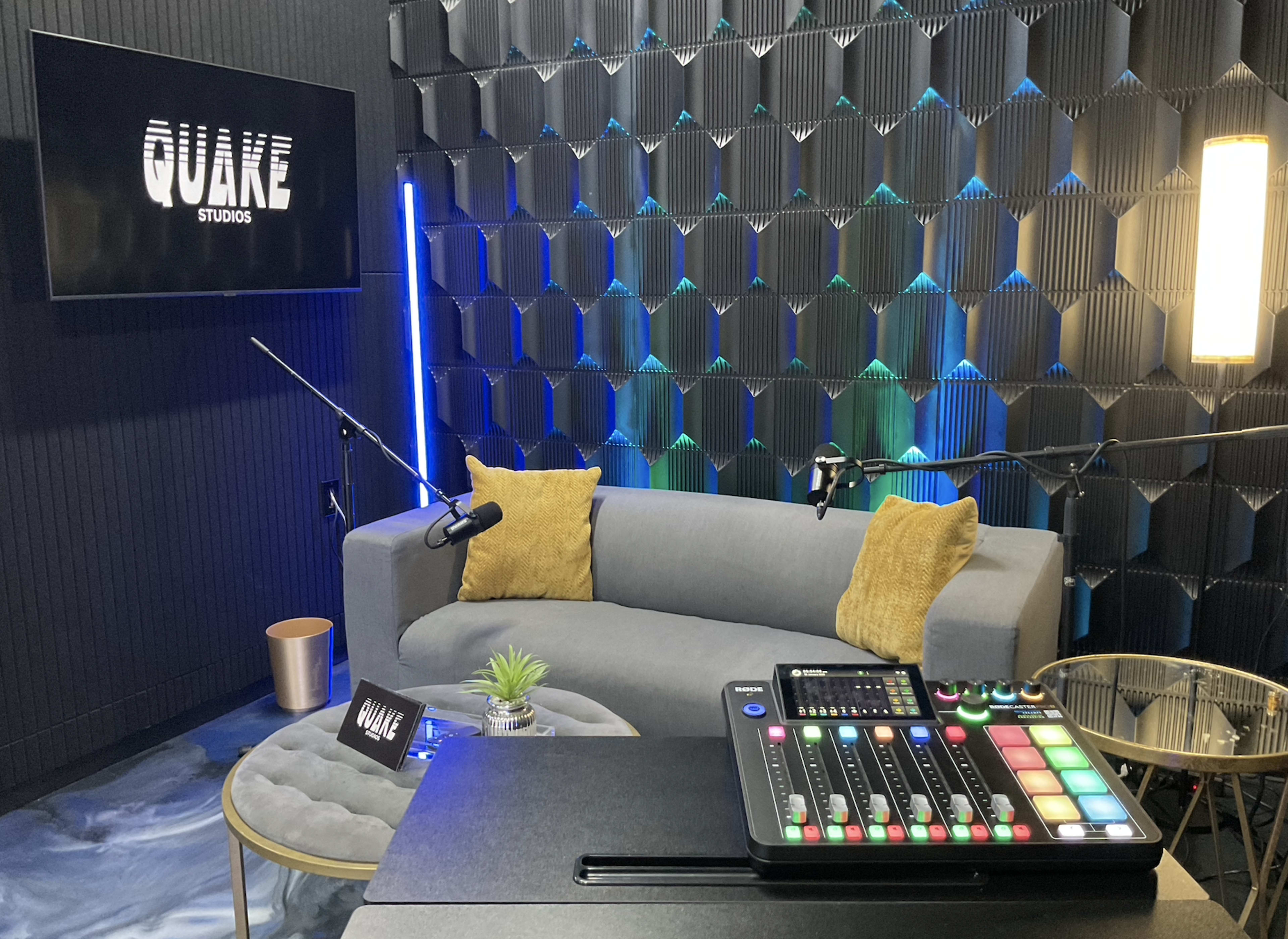 The image shows a modern recording studio featuring a gray couch with yellow throw pillows, a small coffee table, and a control panel with colorful buttons and sliders.