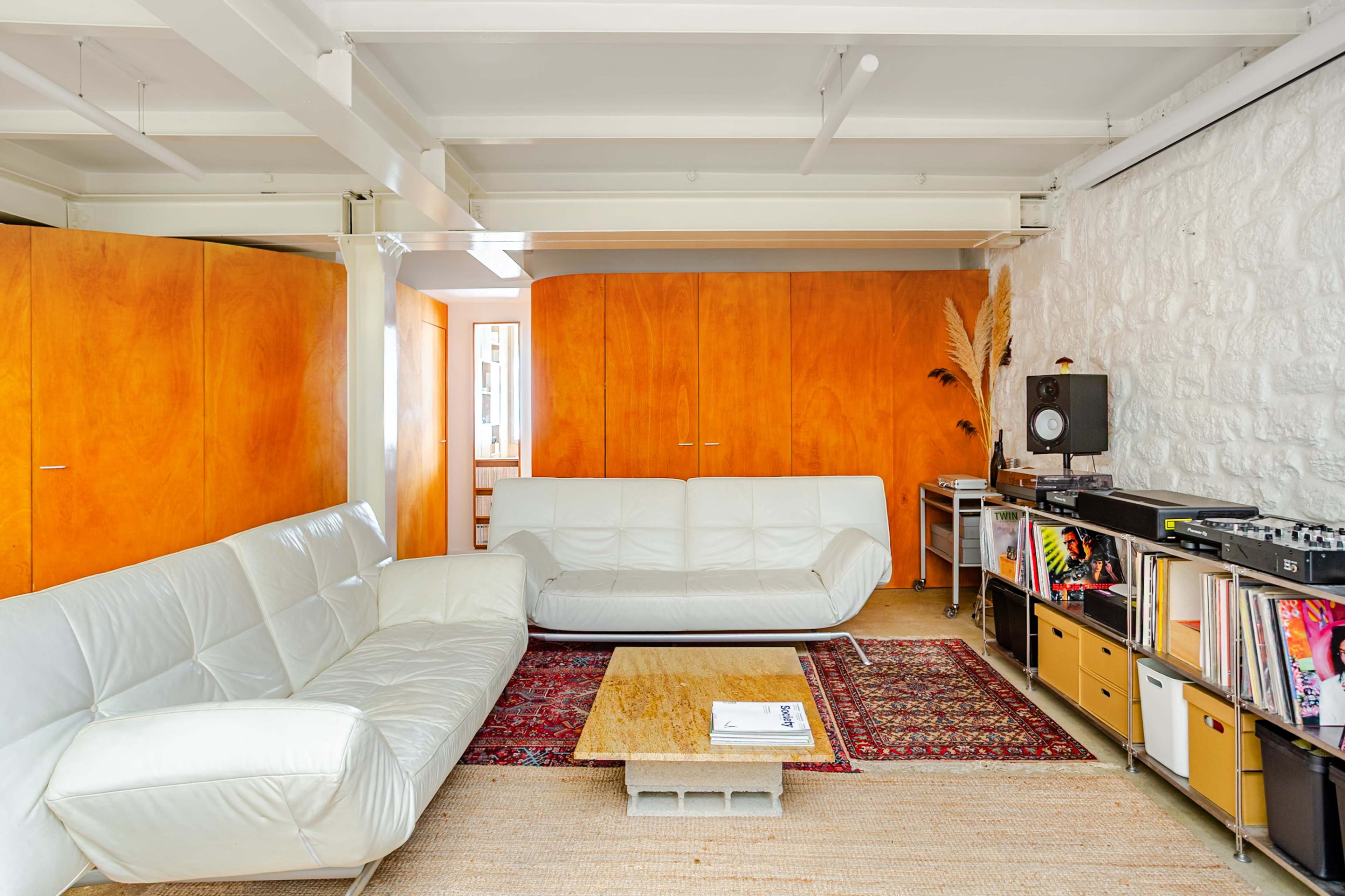 The room features white leather sofas facing a coffee table, with orange wooden cabinets on one wall and a record player with vinyl records on a stand.