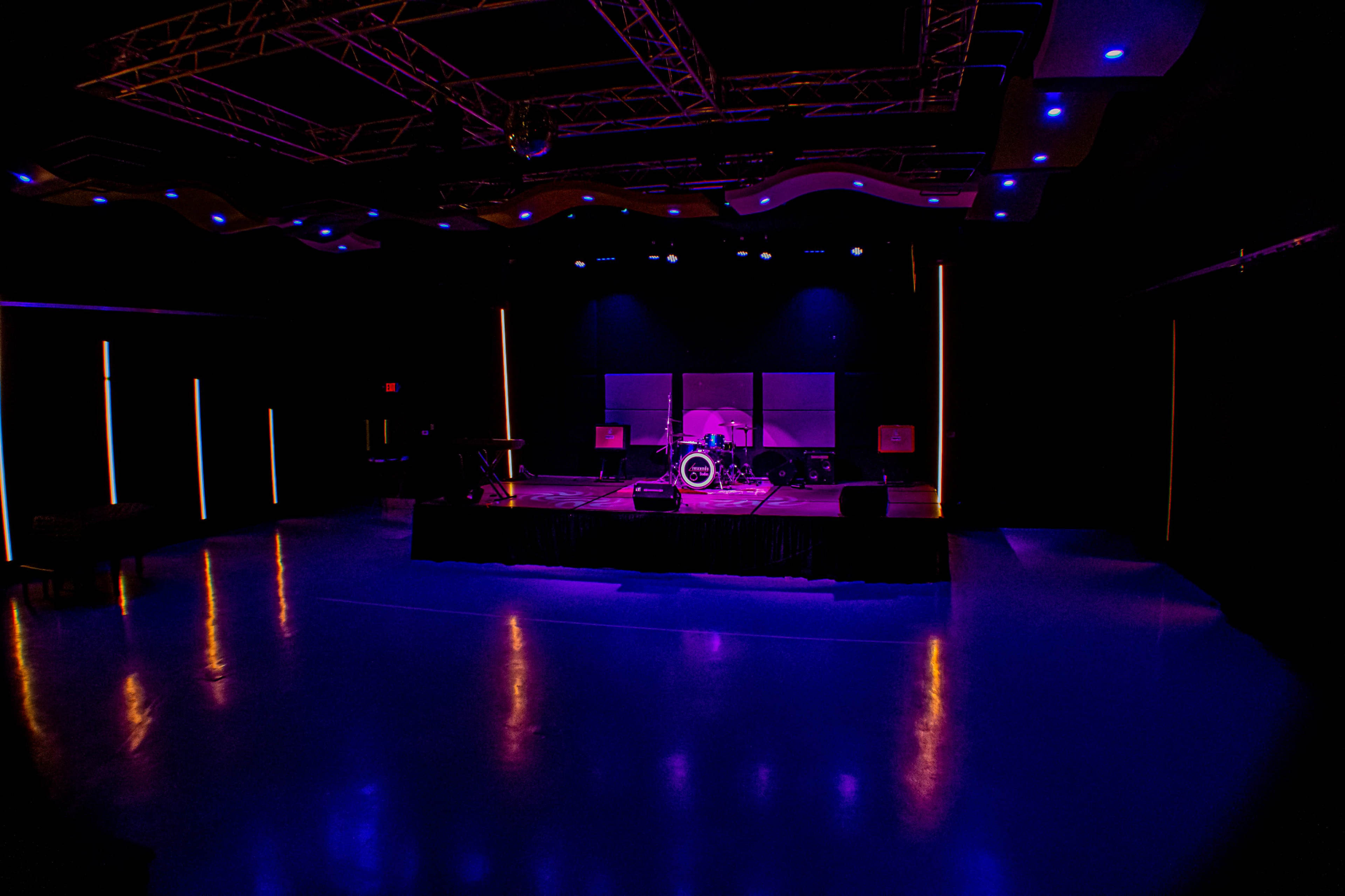A dimly lit stage set up for a performance, featuring a drum kit and various musical instruments with illuminated background panels.