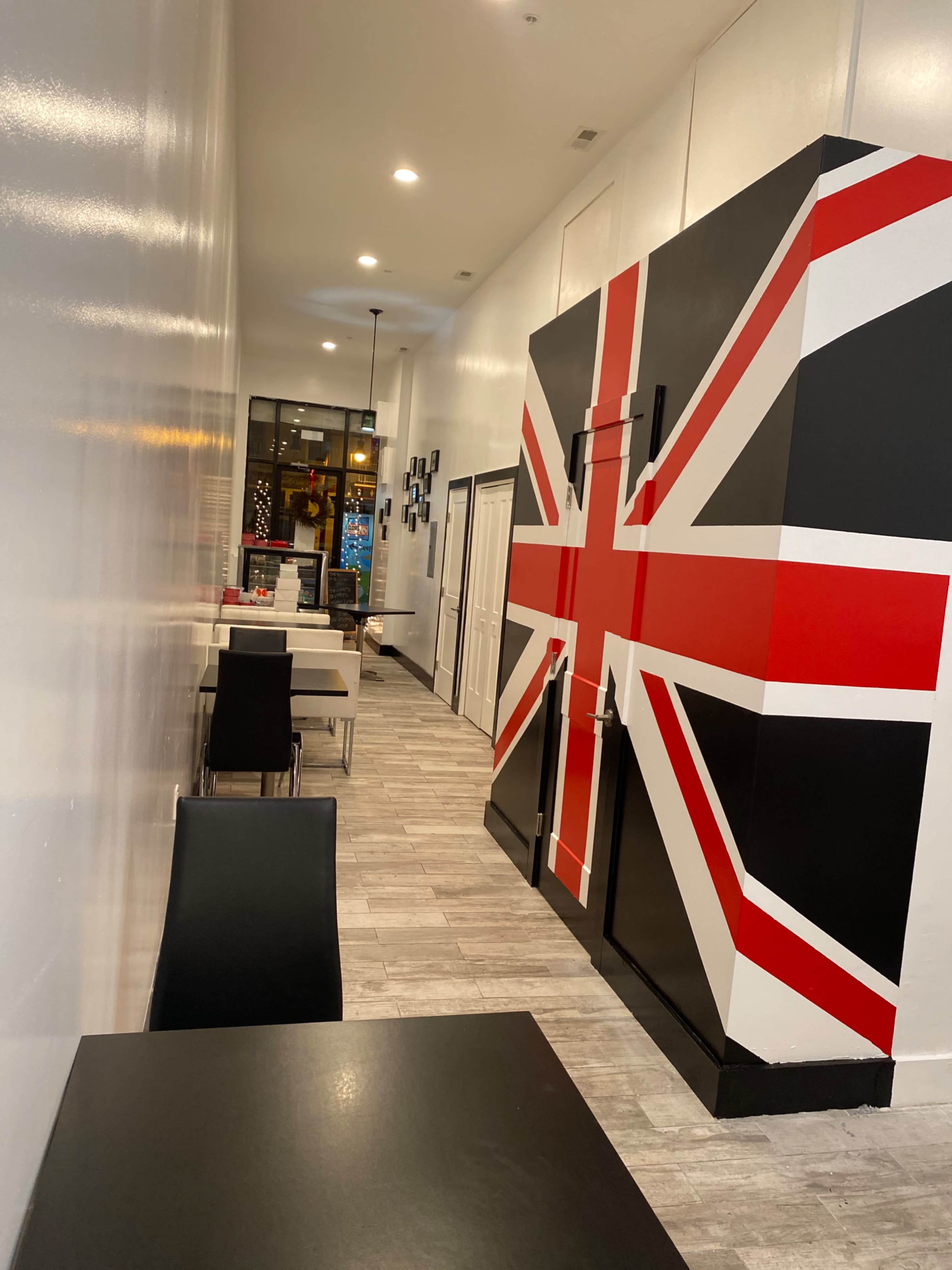 A narrow hallway of a restaurant featuring a large refrigerator with a British flag design and black tables with chairs.