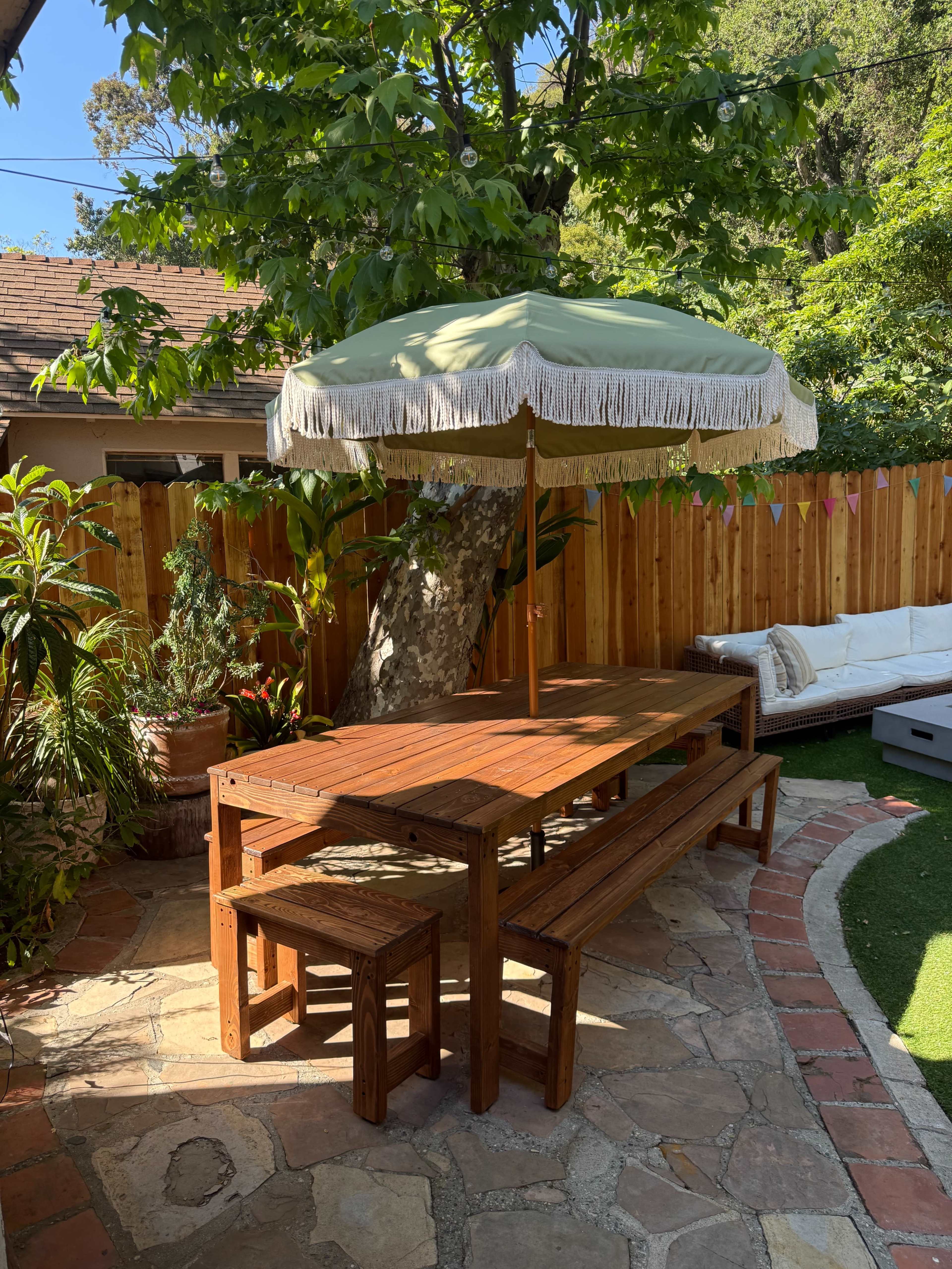 A wooden picnic table with matching stools sits under a green and white fringed umbrella in a landscaped outdoor area.