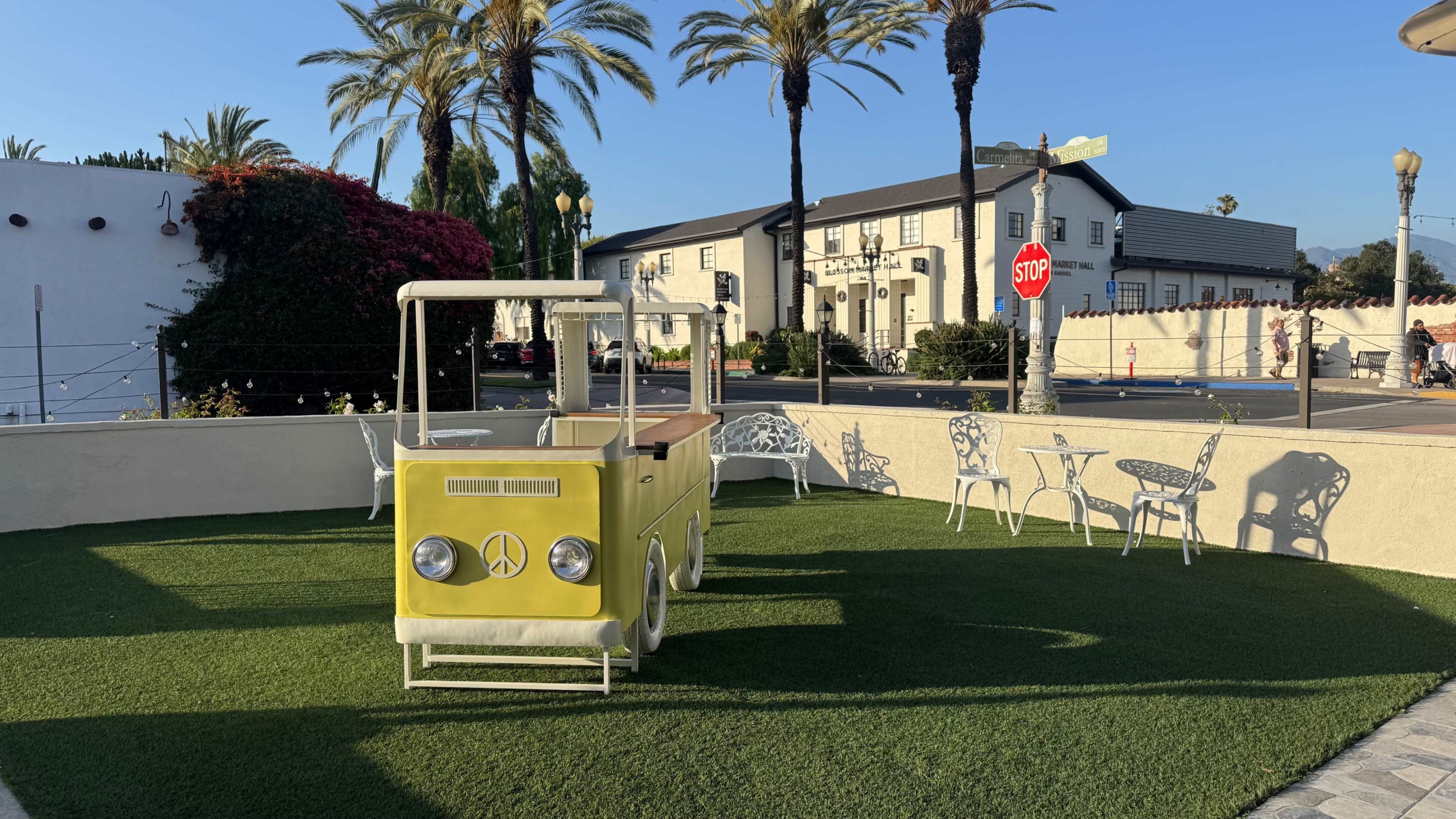 A yellow cart resembling a vintage vehicle is situated on a patch of green grass surrounded by palm trees and outdoor furniture.