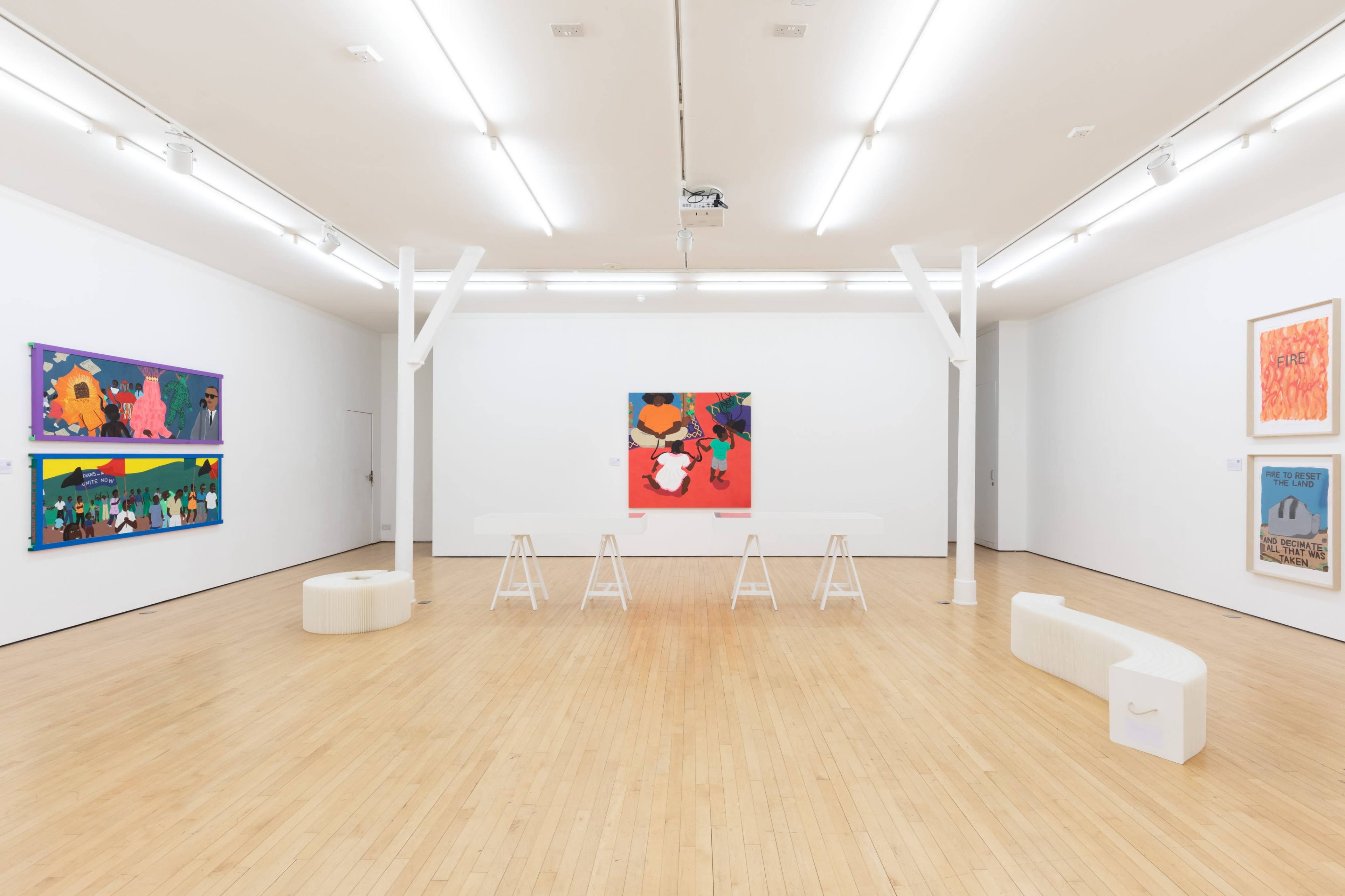 The image shows a modern art gallery featuring several colorful paintings on white walls, with four wooden stools arranged in the center and a few benches along the sides.