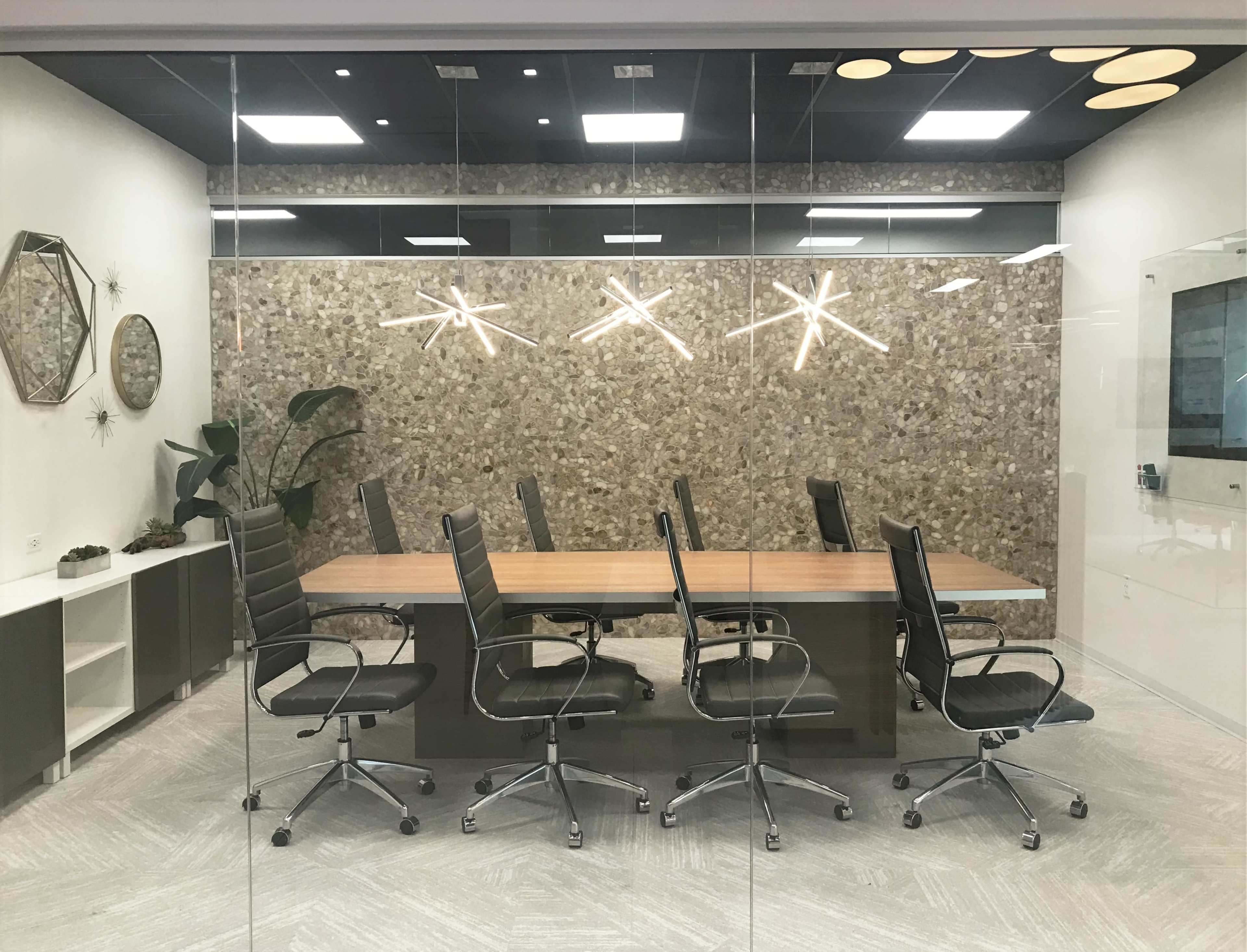 The image shows a modern conference room with a long wooden table surrounded by six black executive chairs, enclosed by a glass wall.