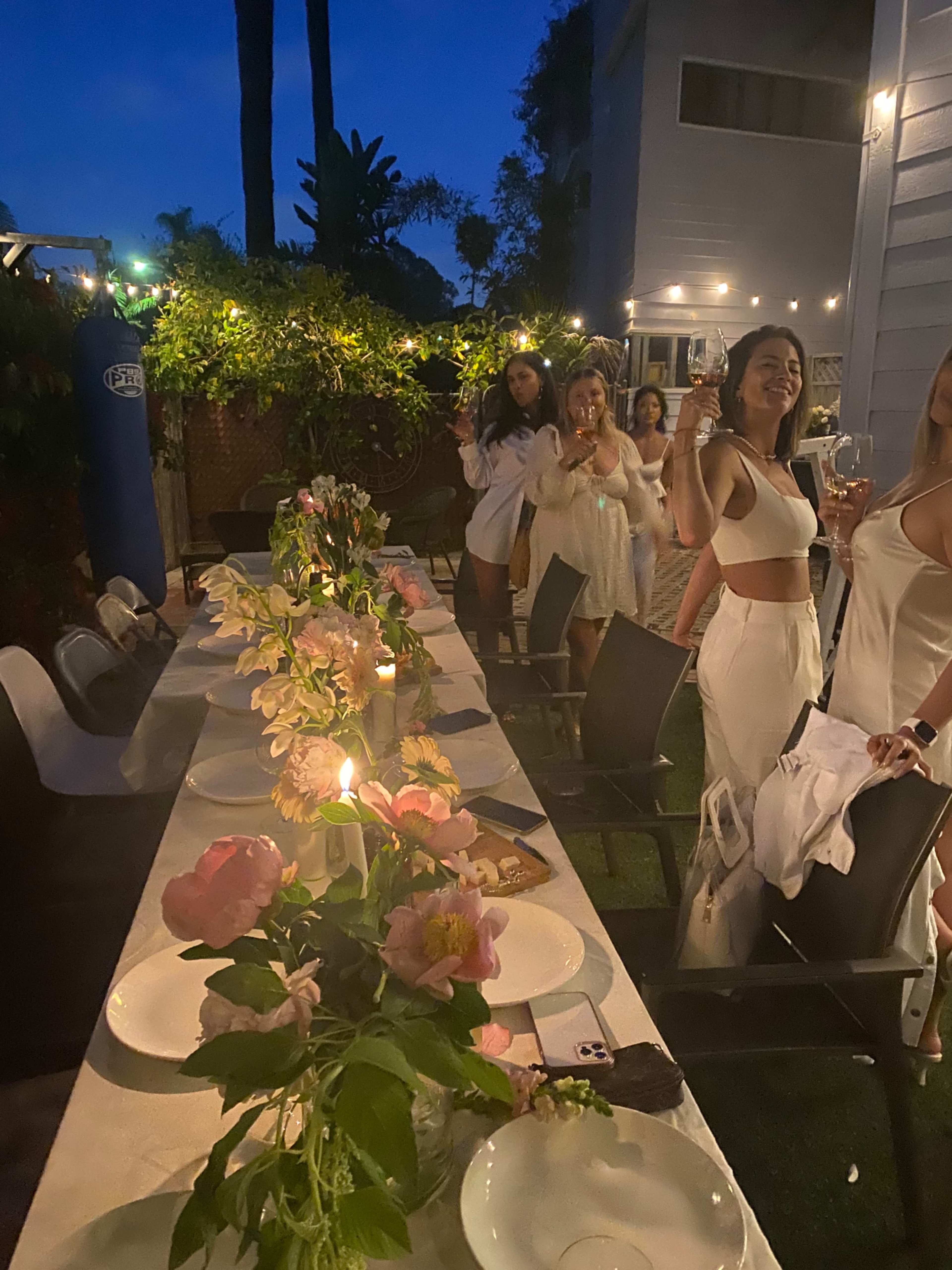 A group of people in white attire raises glasses in a garden setting during a nighttime dinner party, with a long table adorned with flowers and candles.